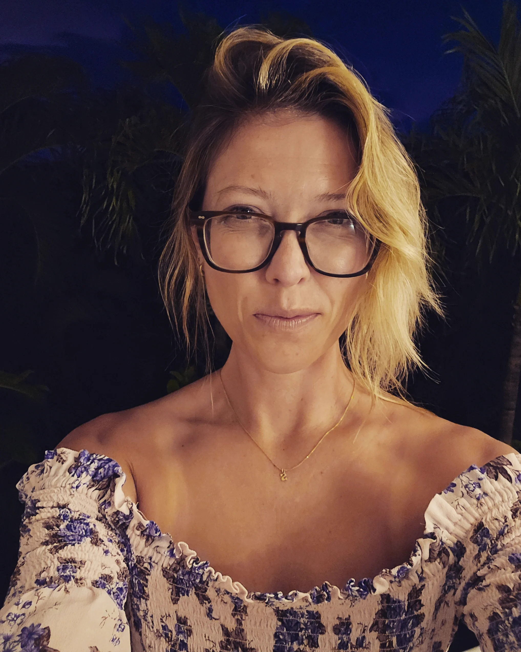 A woman with blonde hair, glasses, and a floral off-shoulder dress is taking a selfie. She is wearing a gold necklace with a small pendant. The background includes dark foliage and a dark blue sky.