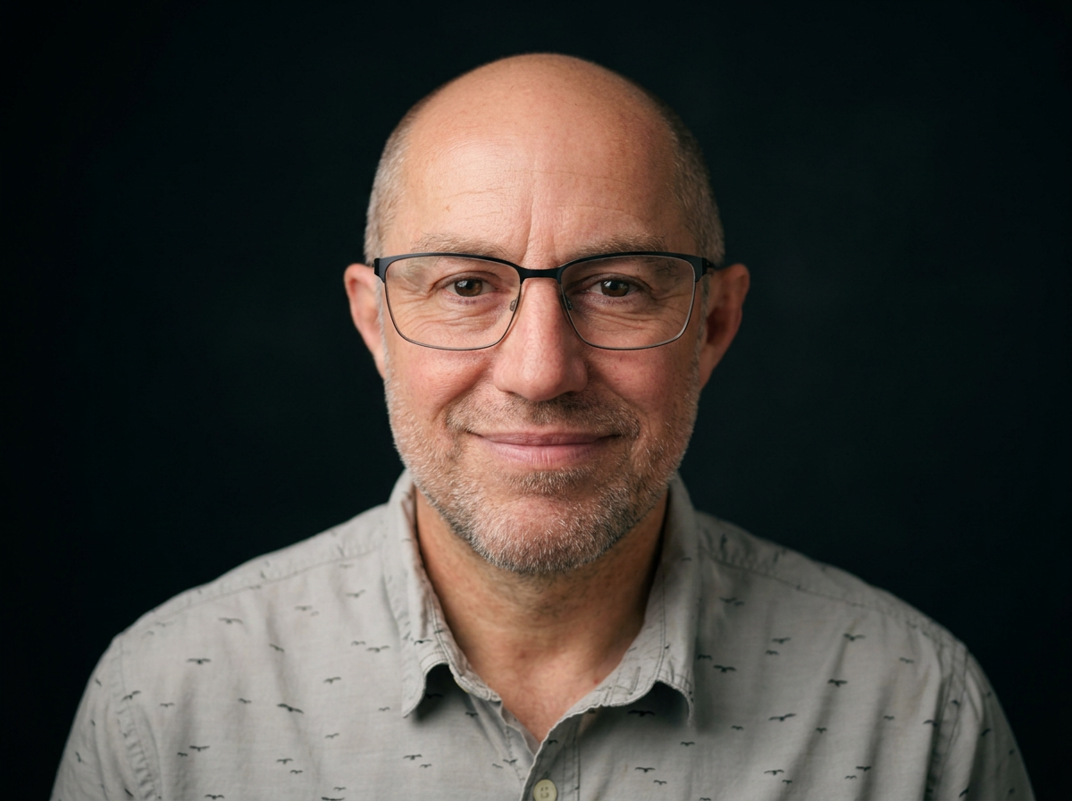 Close-up portrait of a smiling middle-aged man with glasses, short gray beard, wearing a light-colored shirt with small pattern, against a black background.