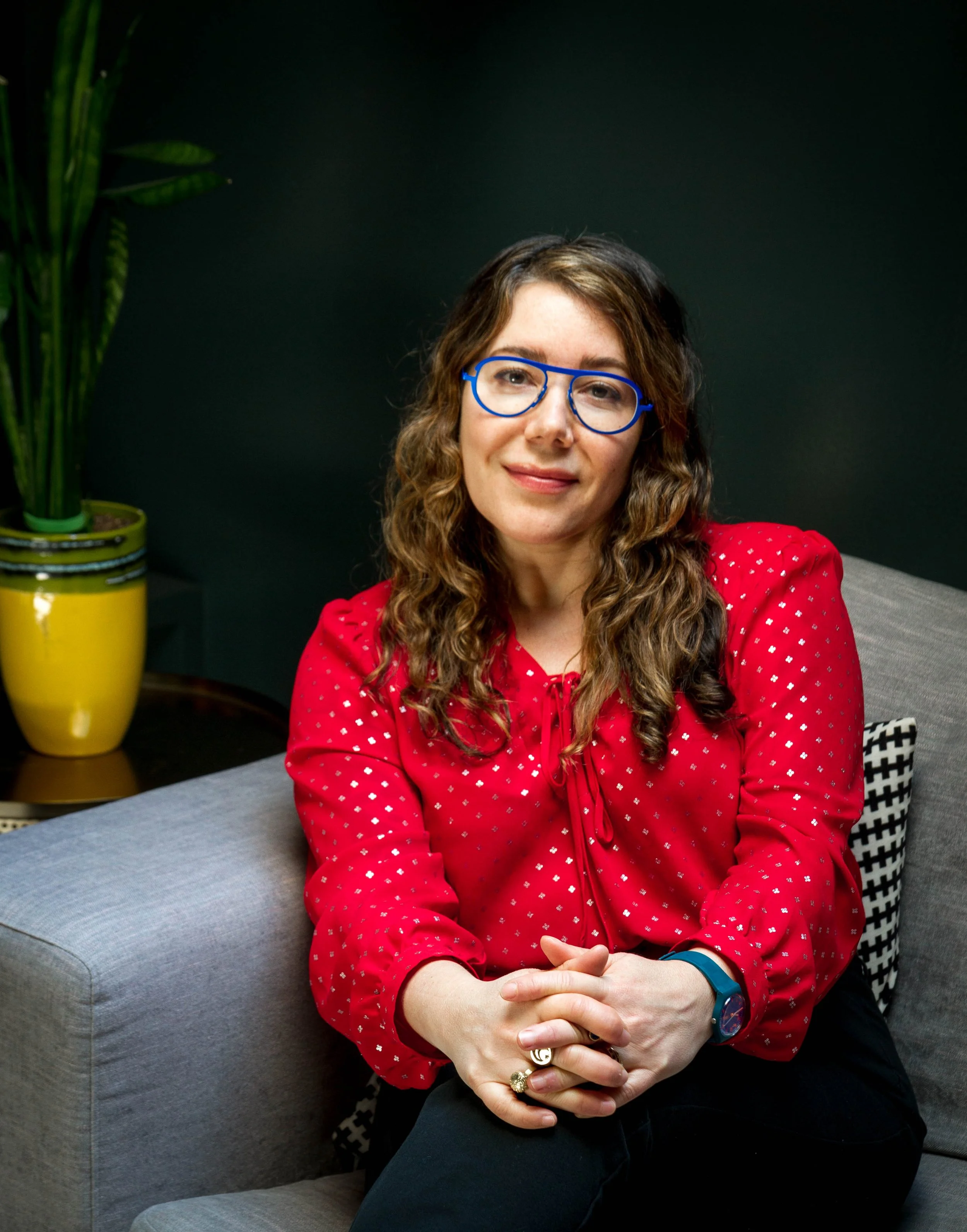 A woman with curly brown hair, wearing glasses and a red polka dot blouse, sitting on a gray couch with a black and white patterned pillow, and a yellow plant pot in the background.