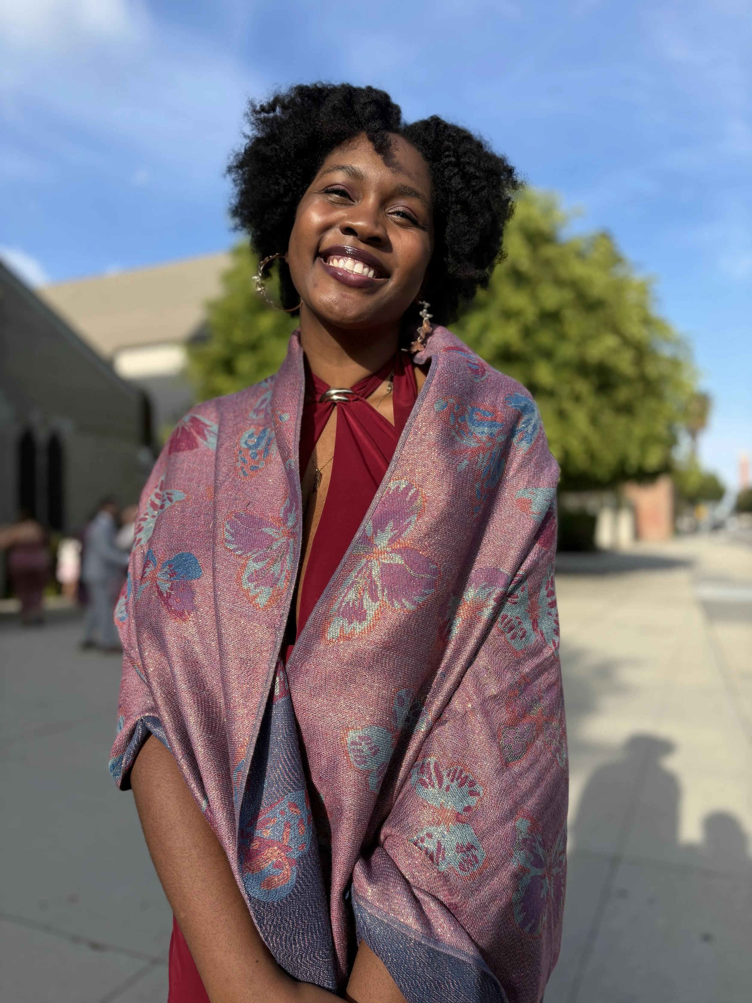 A young woman with curly black hair smiling outdoors on a sunny day, wearing a pink patterned shawl and maroon top, with a background of a blue sky, green tree, and buildings.