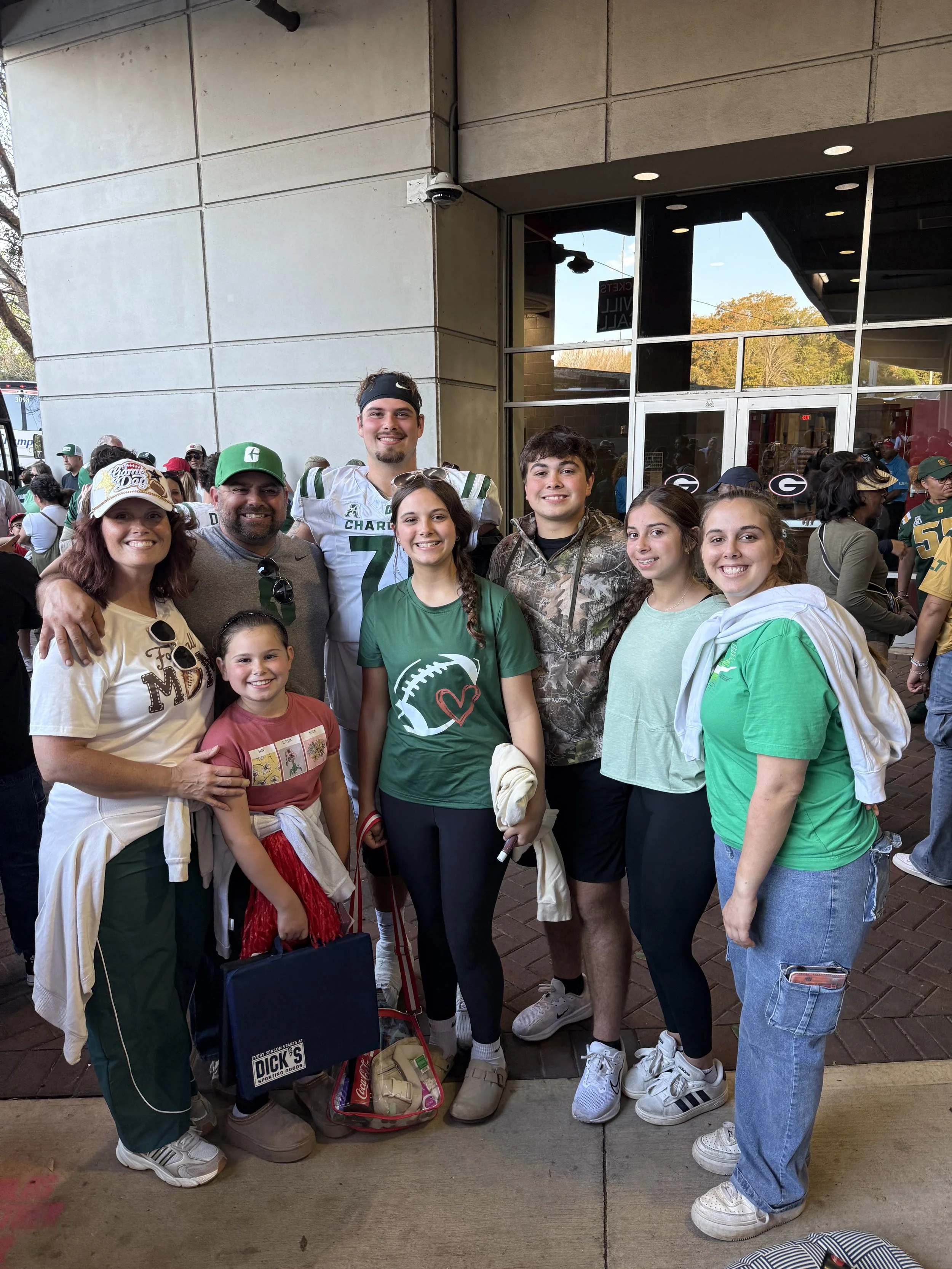 A group of people, including children and adults, standing outside a building with large glass windows, some wearing green and white sports jerseys and caps, smiling at the camera.
