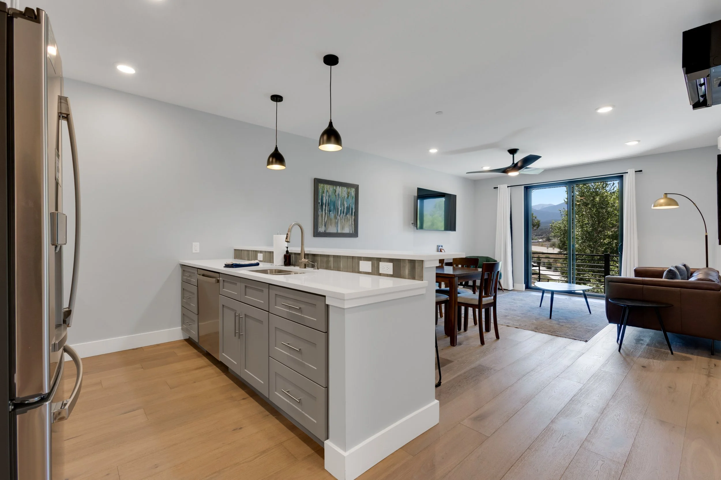 A modern open-concept living space with a kitchen and sitting area. The kitchen features a gray island with a sink and faucet, pendant lights, and hardwood floors. The sitting area includes a wall-mounted TV, a sofa, a floor lamp, and sliding glass doors with white curtains leading to an outdoor balcony with trees and mountains visible in the background.