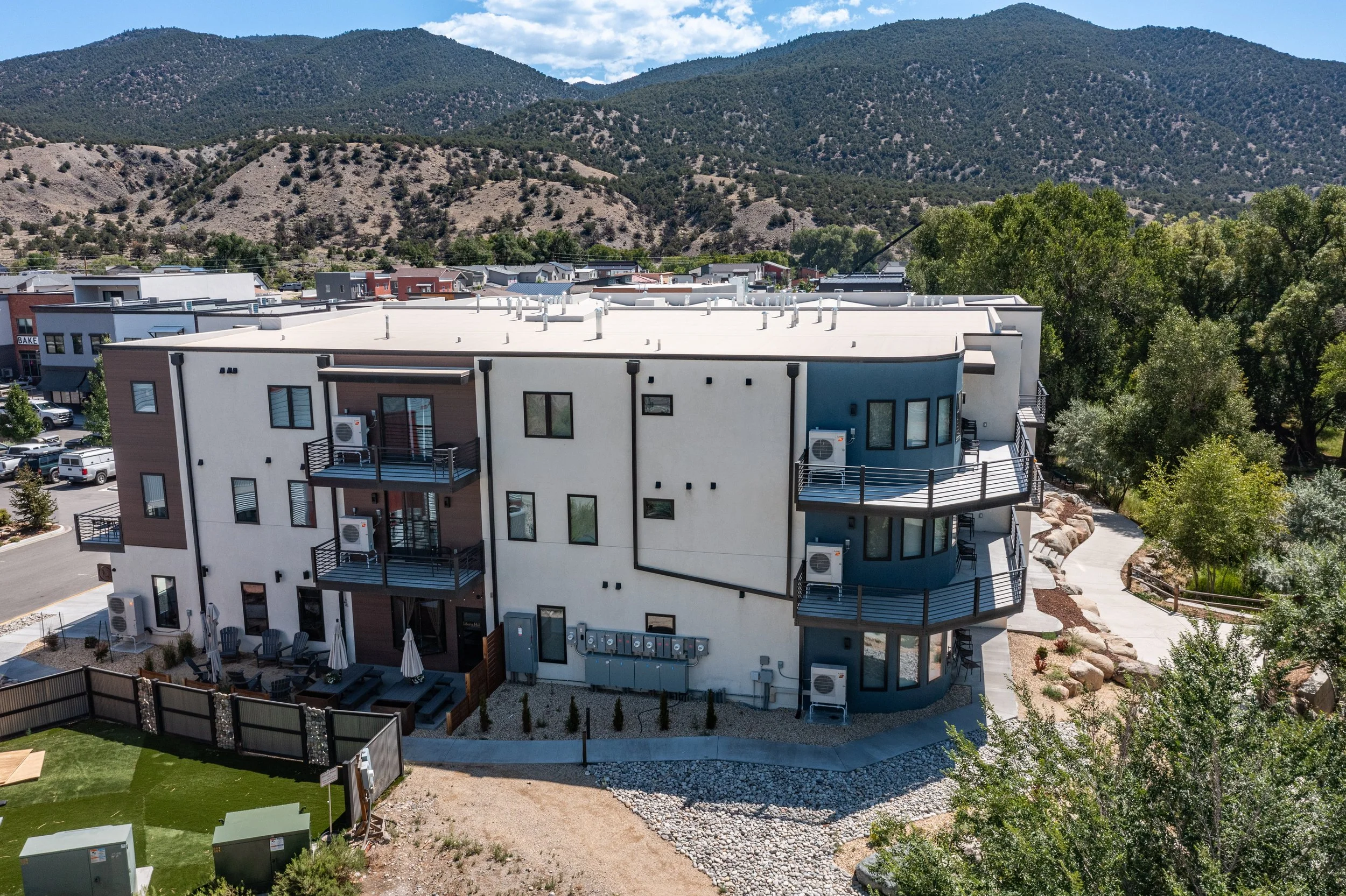 A multi-story residential building with balconies, situated in a scenic area with mountains and trees in the background, under a partly cloudy sky.