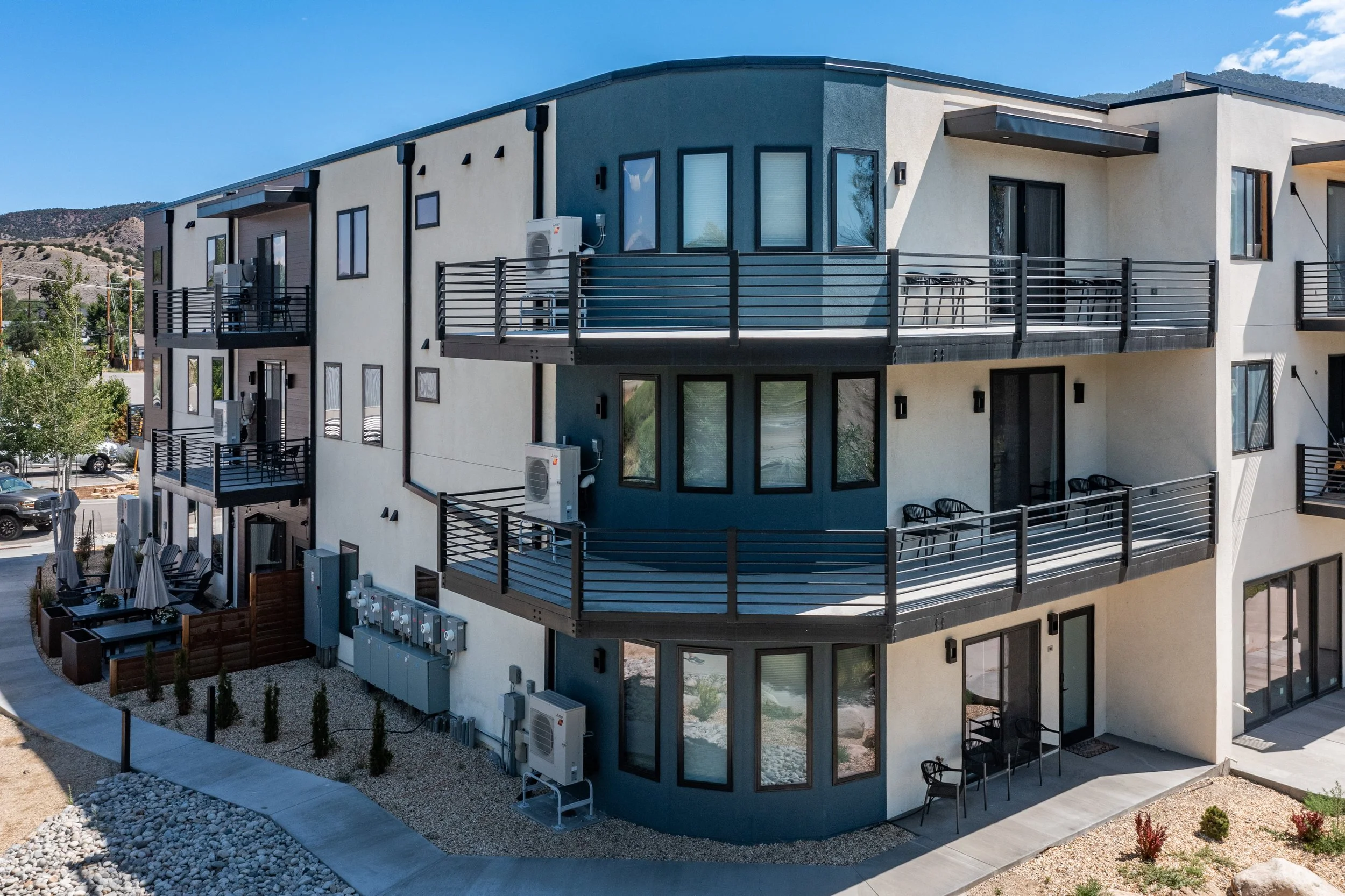 Modern apartment building with three floors, black metal balconies, and large windows, set against a mountain backdrop.