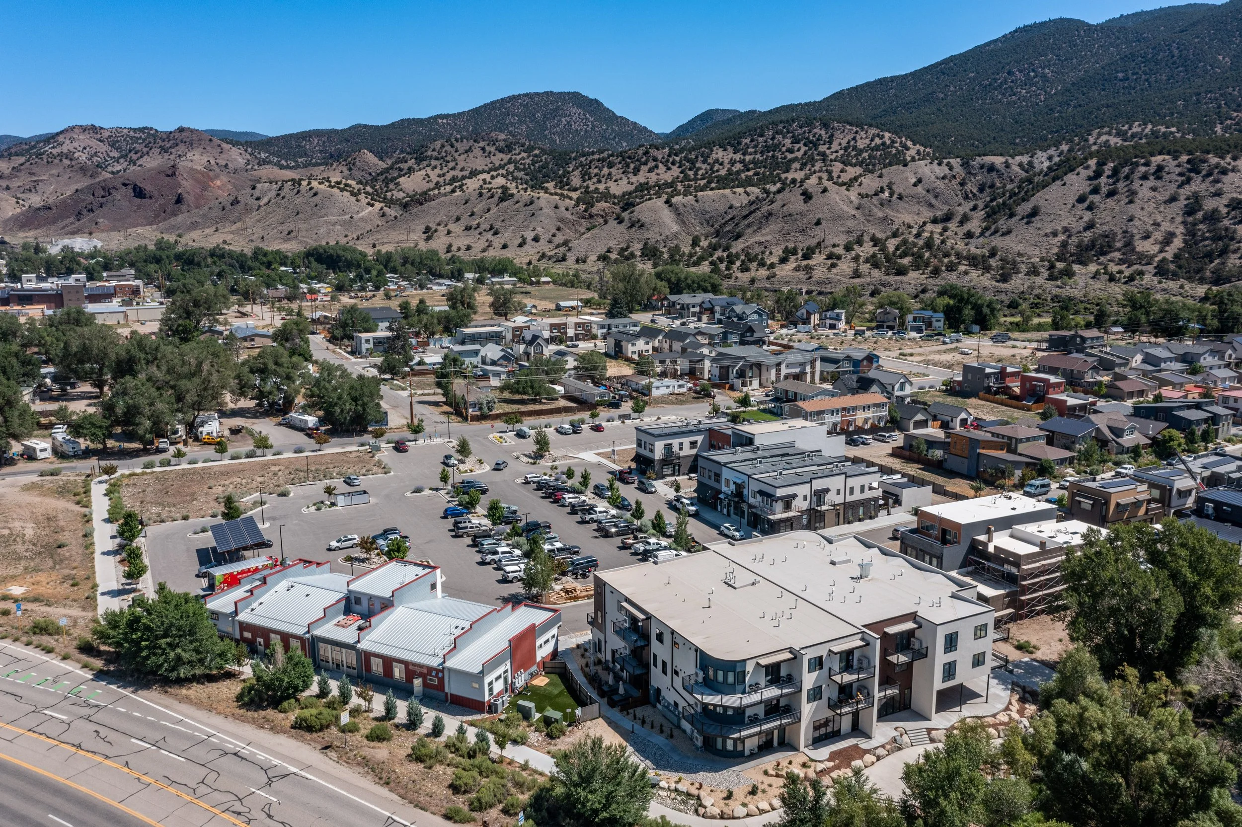 Aerial view of a modern residential neighborhood with apartment buildings, houses, parking lots, trees, and a mountain range in the background under a clear blue sky.