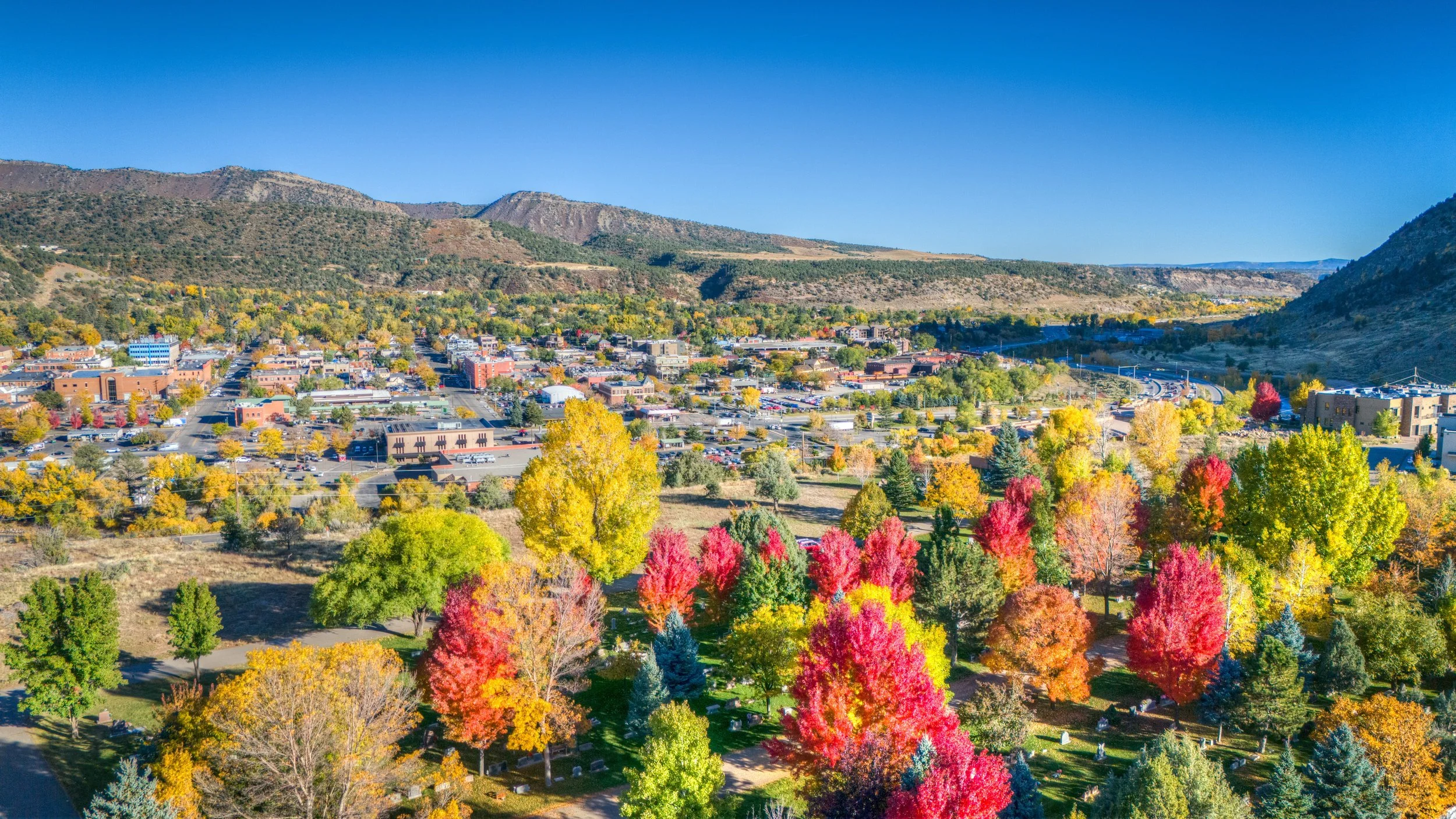 An aerial view of a small town in a valley surrounded by colorful trees and hills under a clear blue sky.