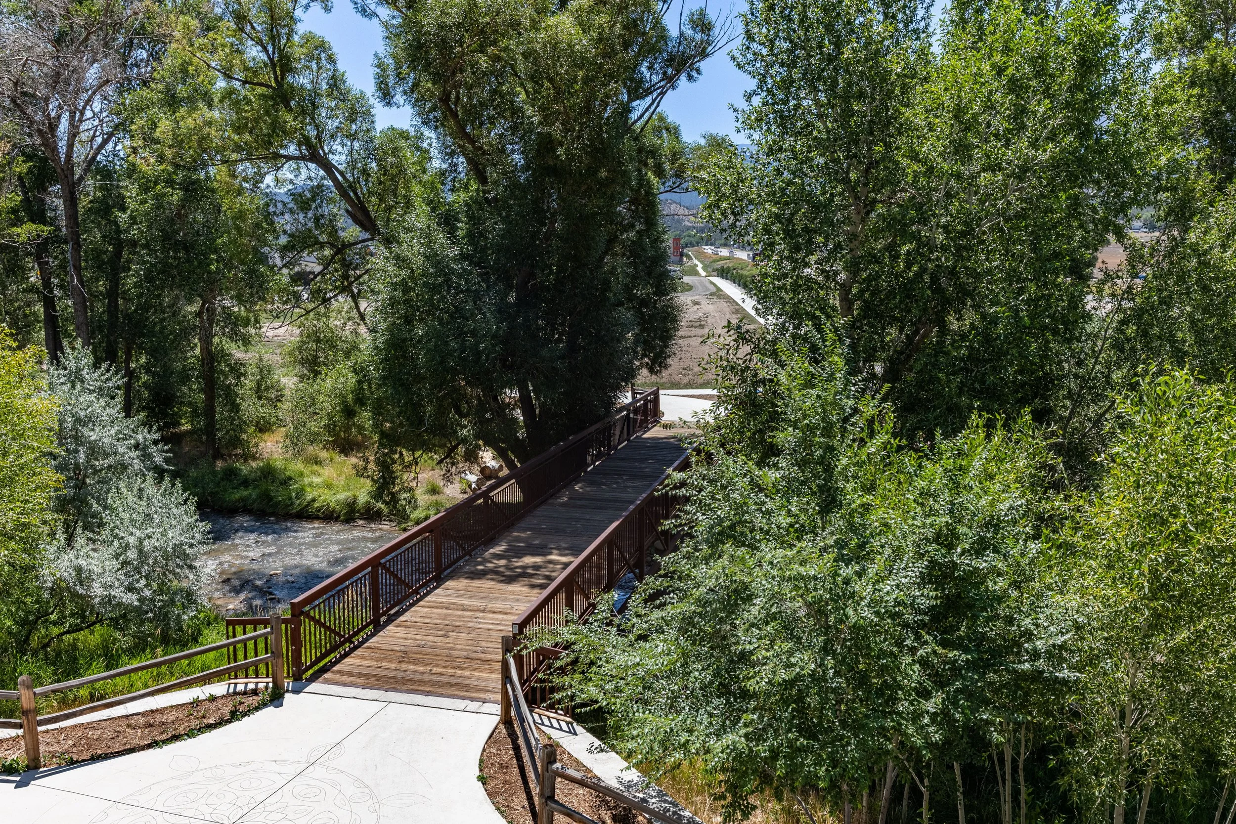 A wooden bridge over a small stream surrounded by large green trees and a pathway leading to buildings in the background.