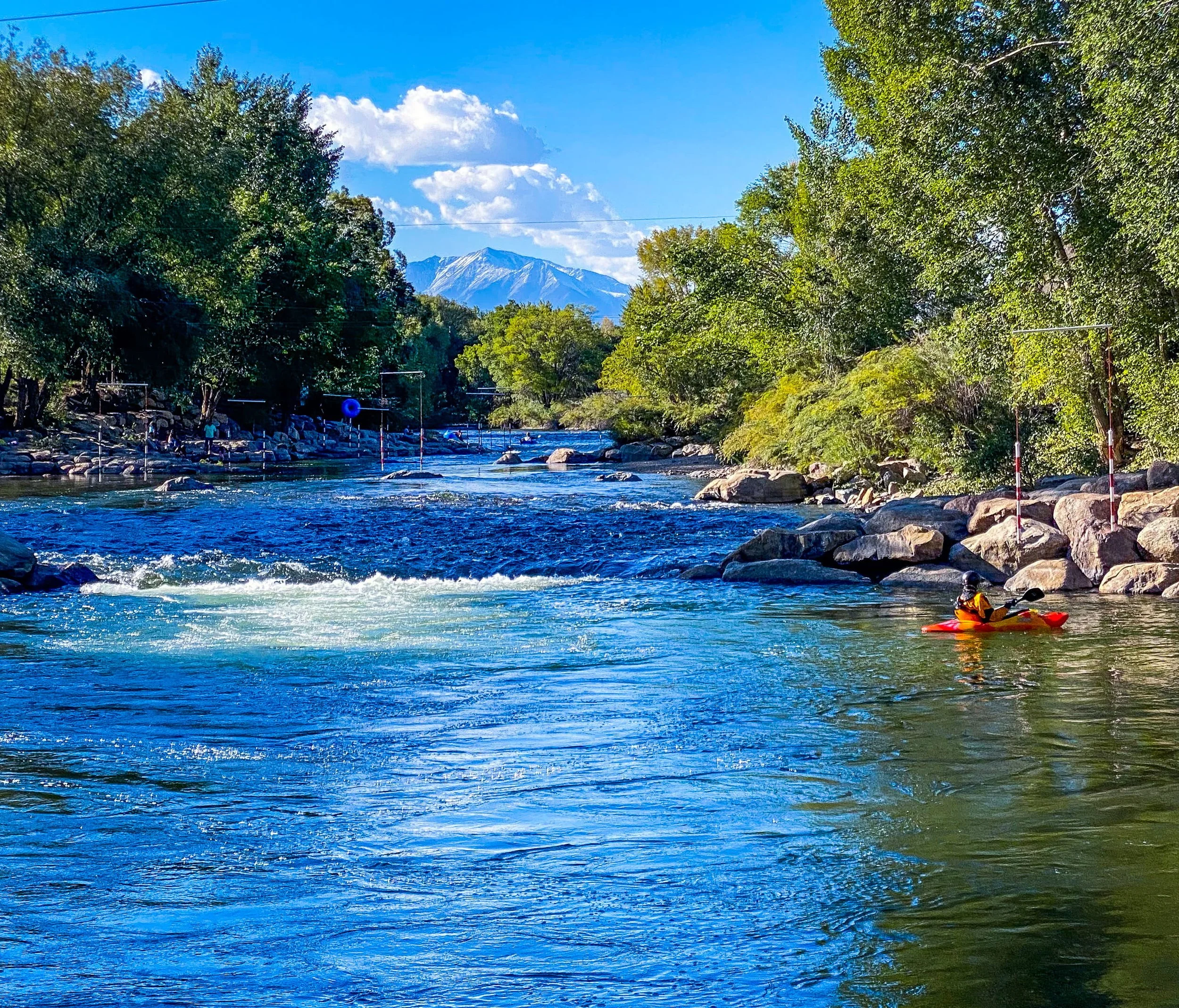 A river flowing through a lush, green landscape with trees on both sides, mountains in the background, and a kayaker in a red kayak near the rocks on the right.