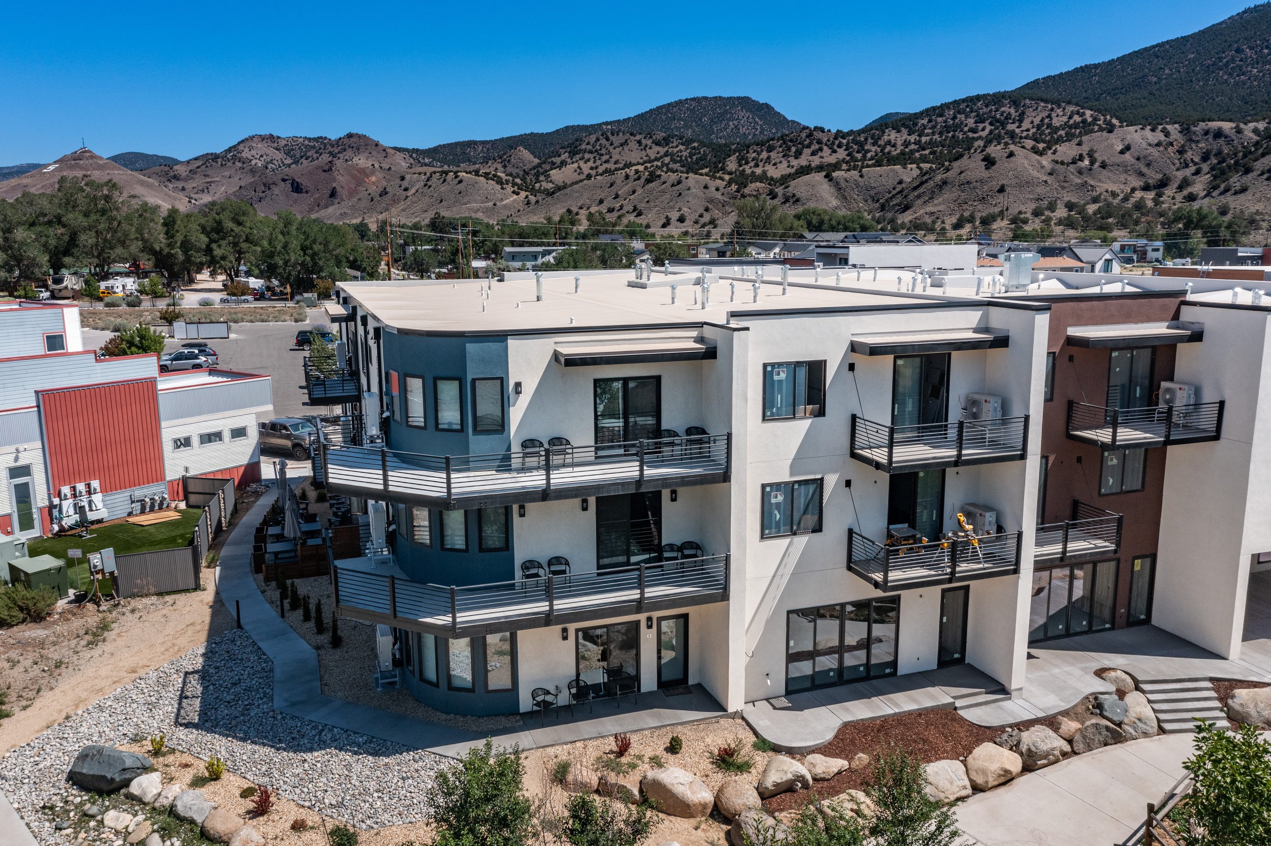 Modern multi-story apartment building with balconies, set against mountains in the background, under a clear blue sky.