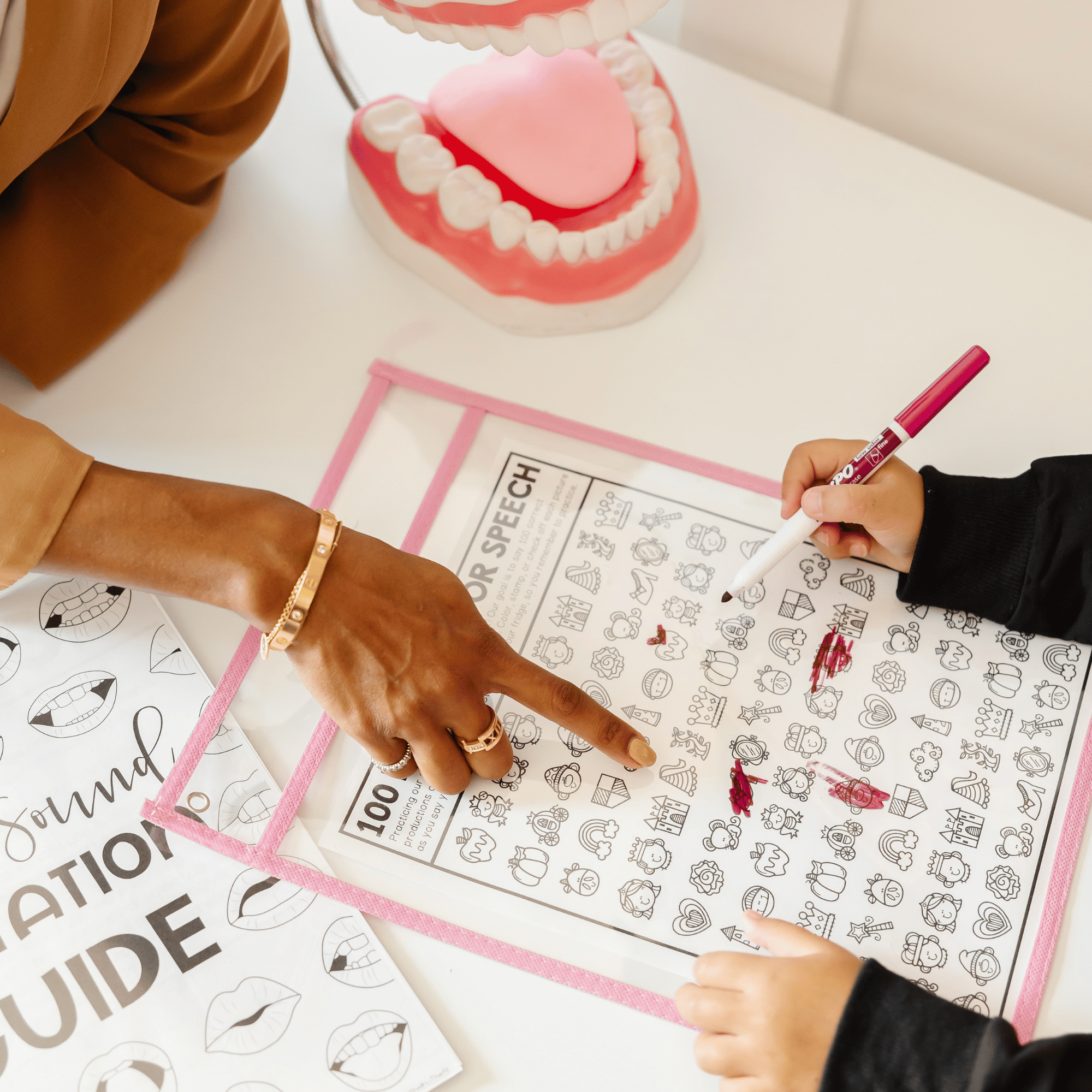 A person using a pink dry erase marker to color a picture of a crown and a happy face on a sheet of coloring pages with various cartoon images, while another person points at the sheet. A dental model with open mouth, teeth, and tongue is in the background on a white table.