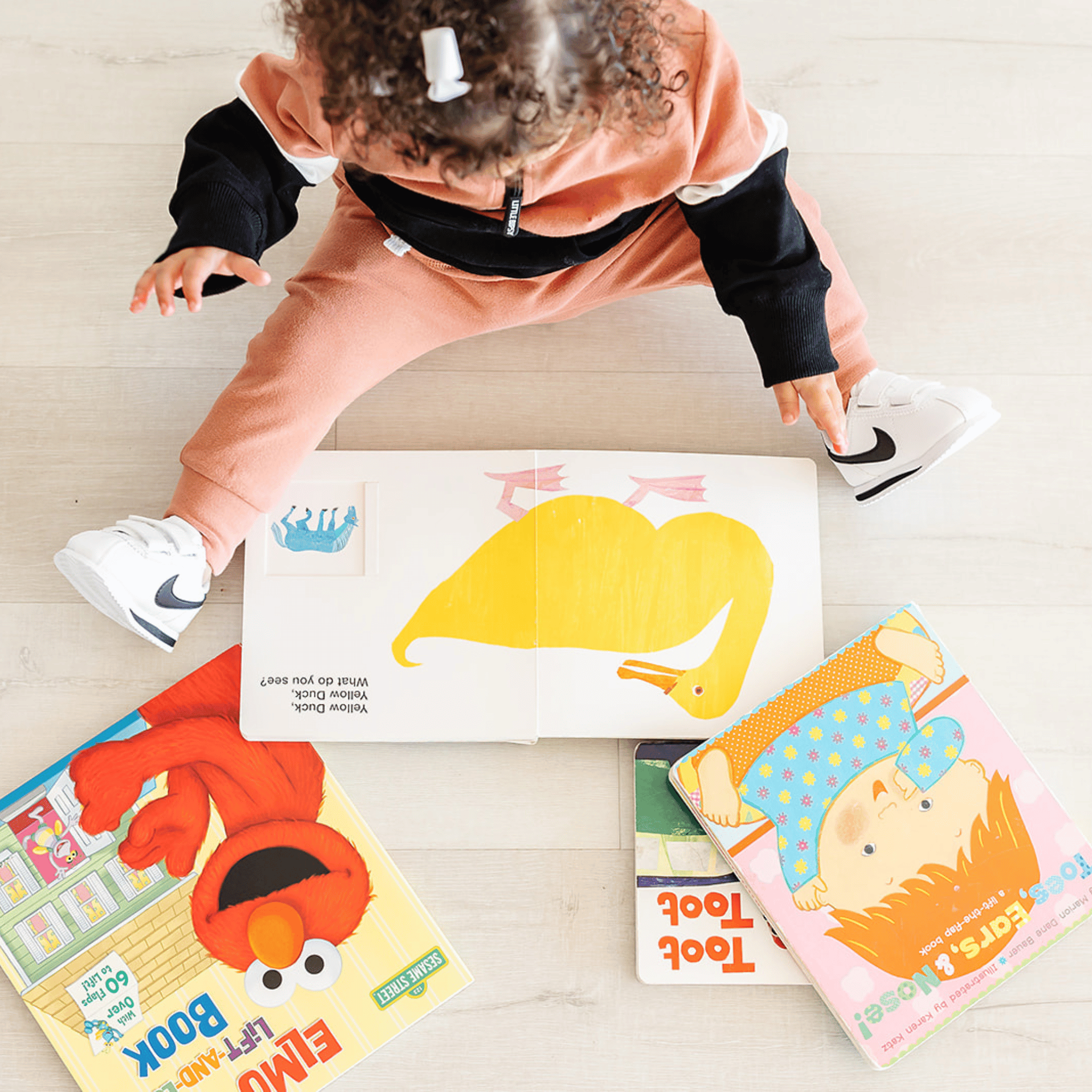 A child dressed in a black and orange costume is sitting on a light-colored wooden floor, surrounded by colorful children's books and a plush toy resembling Elmo from Sesame Street.