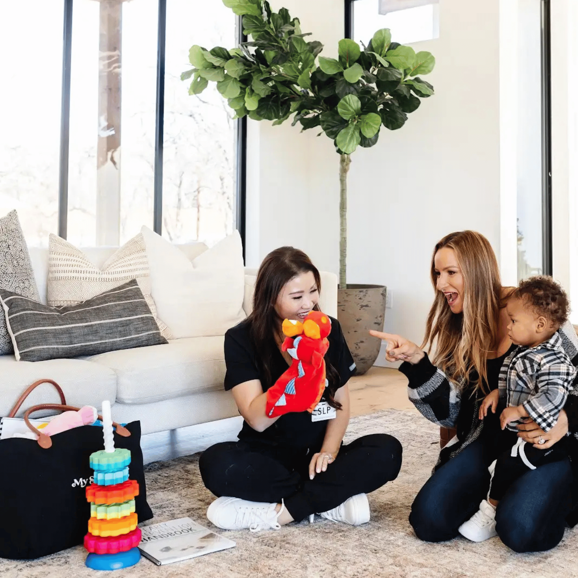 Children playing with stacking toy towers made of colorful interlocking plastic pieces in a room with a large green plant, a sofa with patterned pillows, and a window in the background.