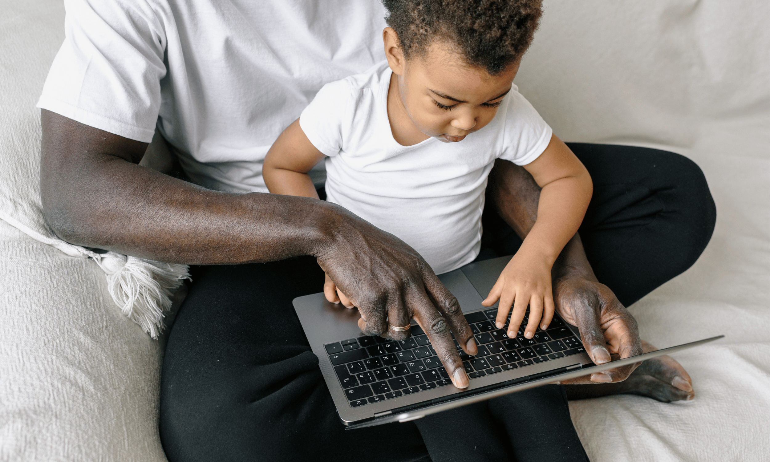 A young child with curly hair and a white shirt sitting on an adult's lap, using a laptop keyboard with adult's hands guiding.
