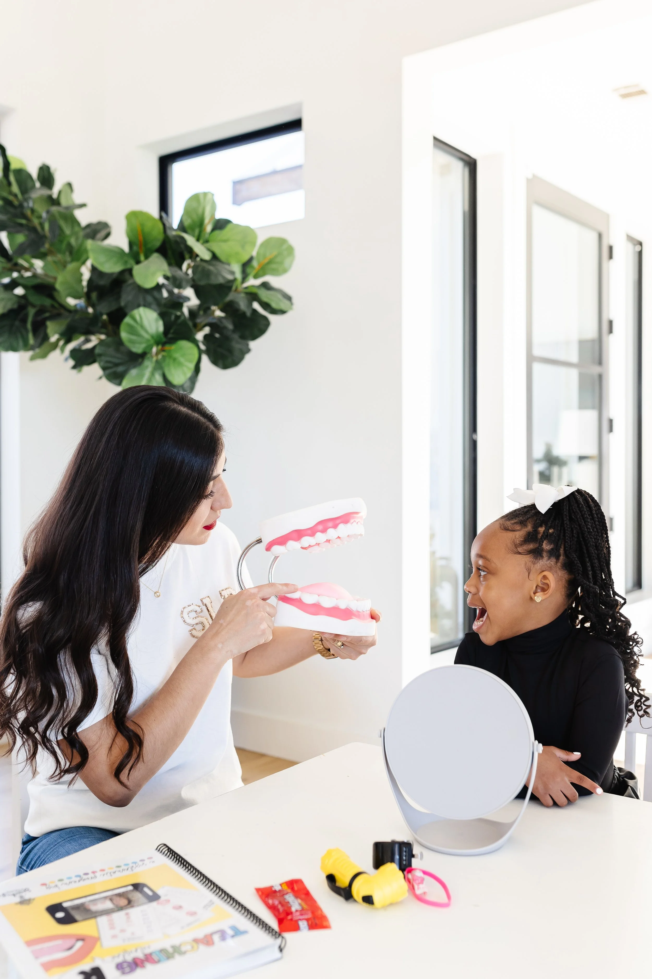 A woman showing a dental model to a young girl at a dental office. The woman has long dark hair and is wearing a white shirt, while the girl has braided hair with a white bow and is wearing a black top. There is a large green plant and windows in the background, and dental tools and a mirror are on the table.