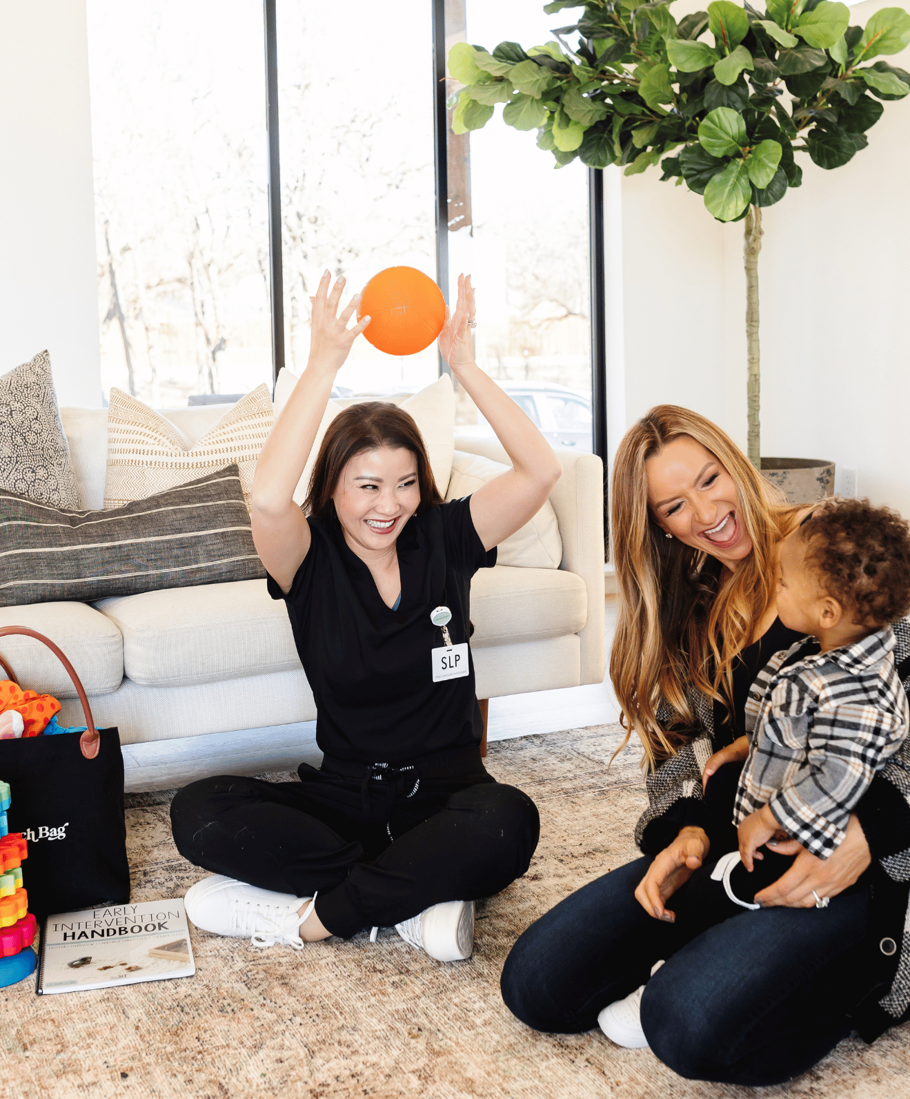 A woman in a black shirt, sitting on the floor and holding an orange ball above her head, smiling. Another woman with long red hair, wearing a black top, crouching and talking to a young girl with curly hair, dressed in a plaid shirt. The room has a beige couch with cushions, a large green plant, and window letting in natural light. There are toys and a book titled 'Early Intervention Handbook' on the floor.
