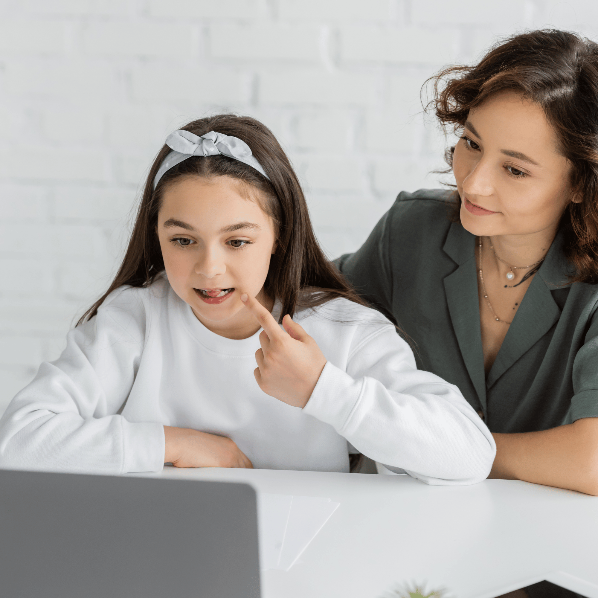 A woman appears to be instructing or explaining something to a young girl, who is pointing at a computer screen. There is a white brick wall in the background.