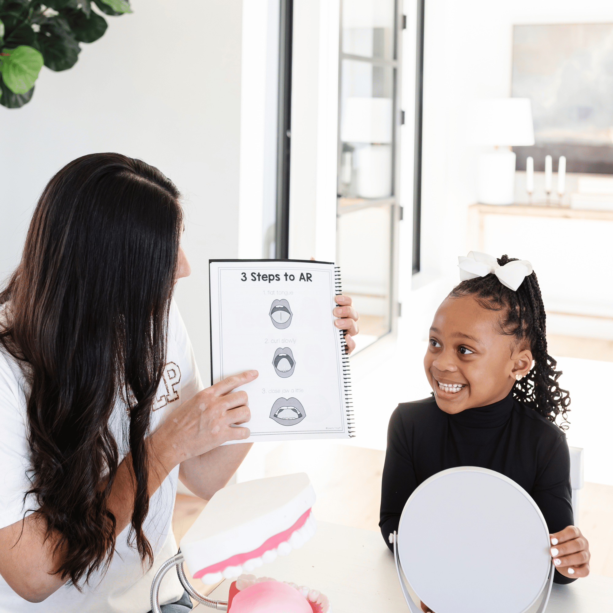 A young girl with black curly hair and a white bow on her head smiling, looking at a woman with long brown hair and tan skin, who is holding a sheet with step-by-step instructions about creating augmented reality experiences with mouth gestures. The woman is pointing at the sheet, and there is a dental model with a pink base and white teeth in the foreground. The background shows a bright, modern room with a white wall, a glass door or window, and some furniture.