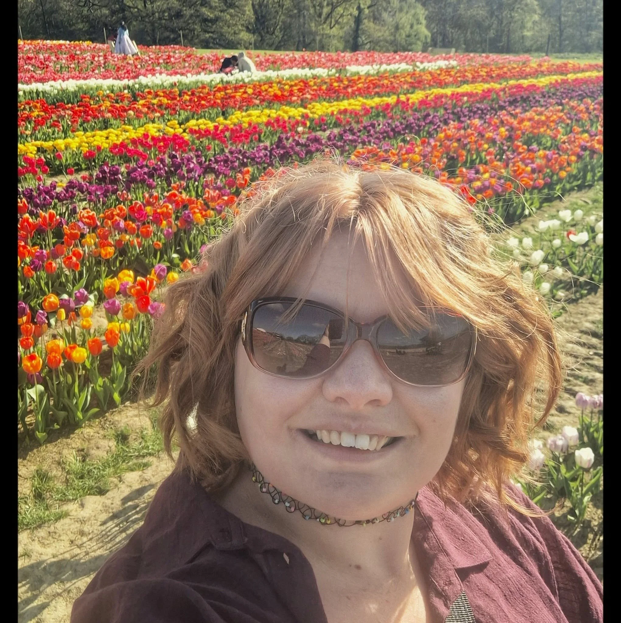 Close-up of a woman's face wearing sunglasses, with a colorful tulip garden in the background.