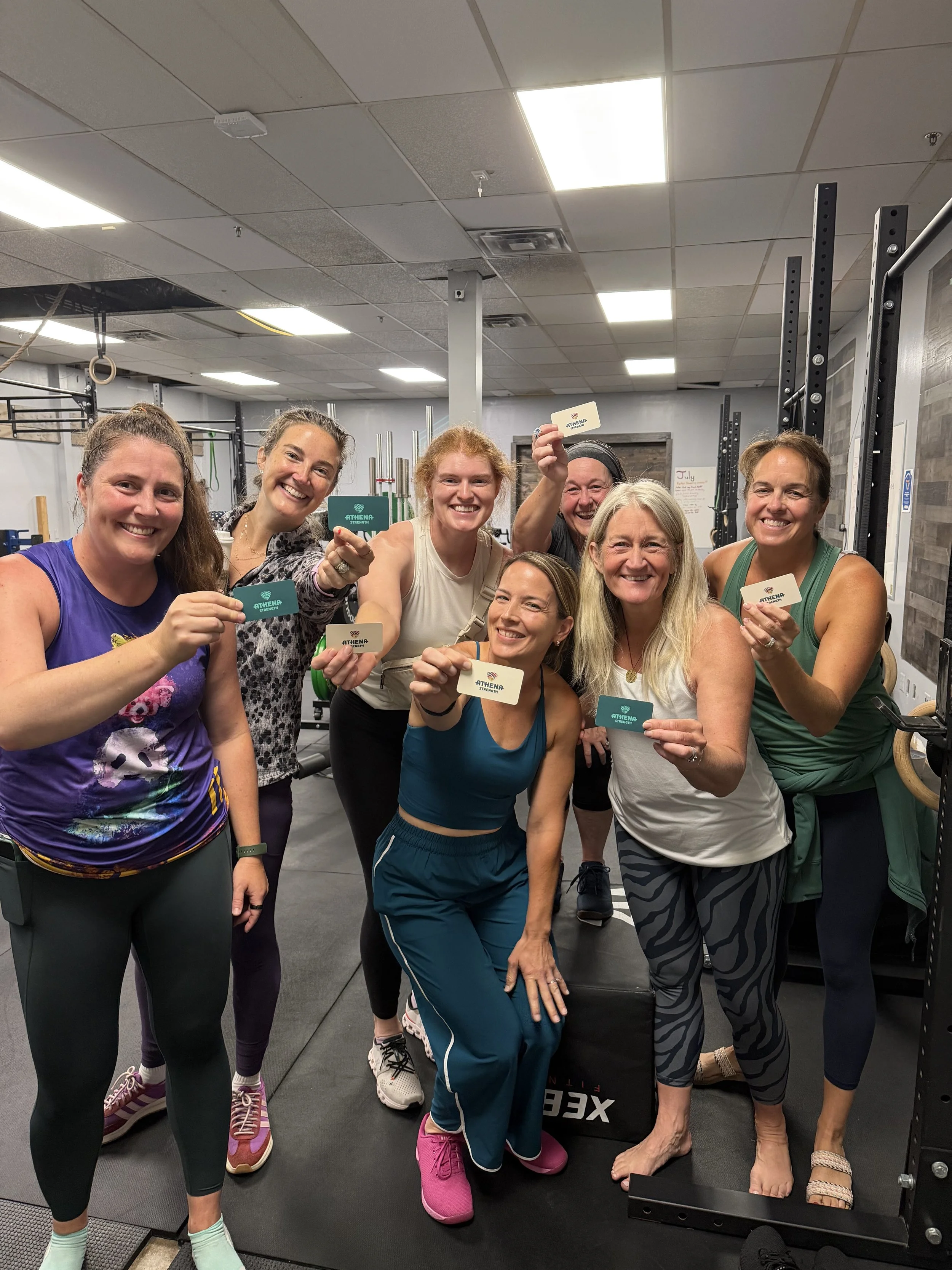 Group of women smiling and holding cards in a gym, with one woman standing barefoot on a platform.