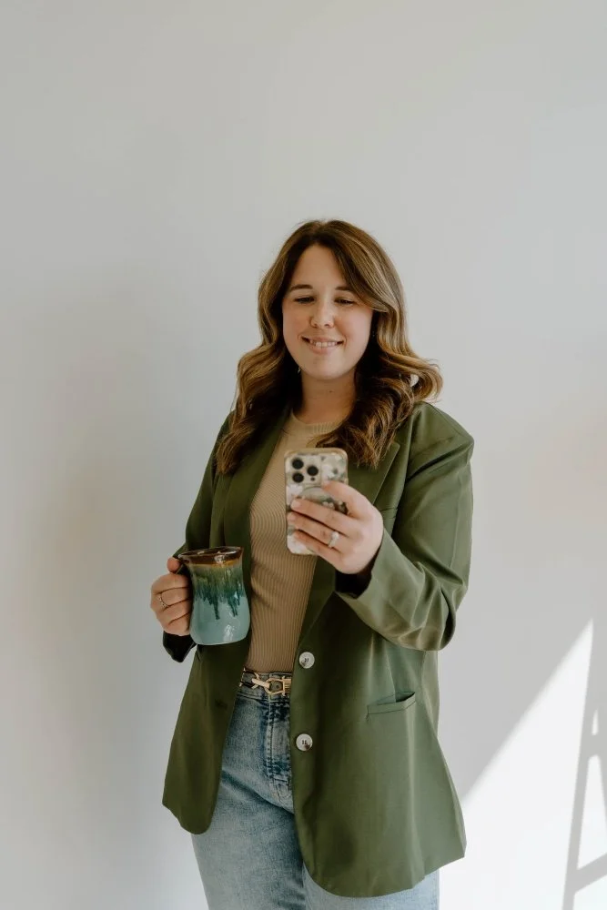 Michella Hands looks at her phone in her hand, holding a teal green mug in the other hand. She has long dark brown hair, fair skin, and wears a dark green blazer, beige shirt, and jeans.