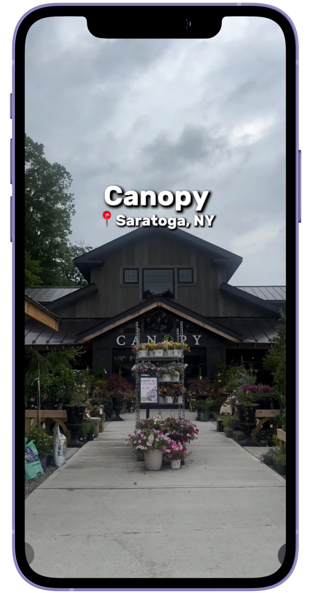 A smartphone screen displays the entrance to a garden center named 'Canopy' in Saratoga, NY. The entrance is surrounded by flowers and potted plants, with a sign in front advertising a sale on flowers. The building has a rustic look with dark wood siding and large windows, set against a cloudy sky.