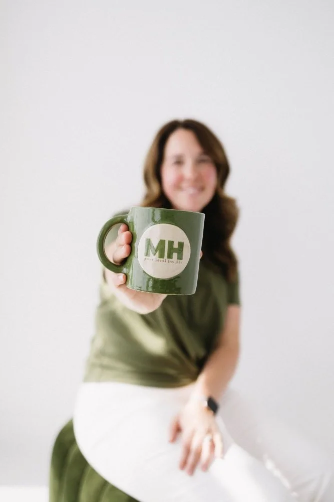 Michella holds a green mug with the letters MH to the camera. The mug is the only thing in focus, with Michella out of focus behind the mug.