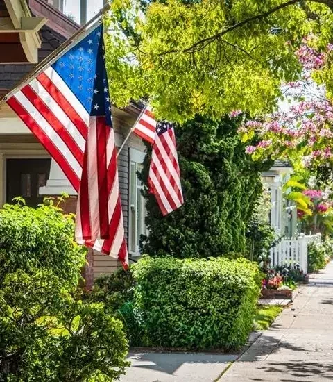 Two American flags hanging outside a house on a porch, surrounded by green bushes and trees with colorful flowers in the neighborhood.