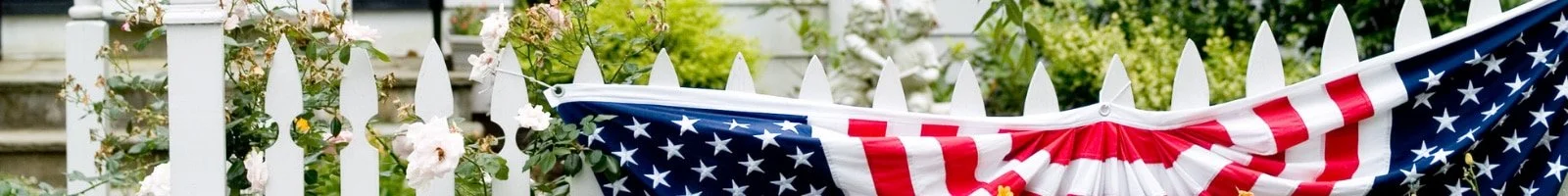 American flag bunting hanging on a white picket fence with flowering plants in the background.