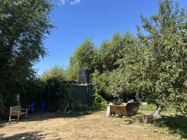A backyard with green trees, a wooden bench, a black water tank on a small shed, and a black fire pit or grill, under a clear blue sky.