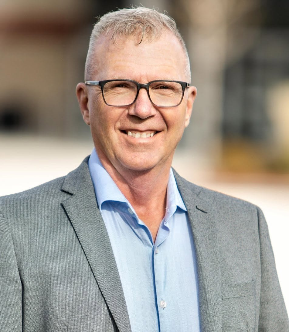 A middle-aged man with short blonde hair, glasses, and a smile, wearing a gray suit jacket and a light blue dress shirt, standing outdoors in natural light.