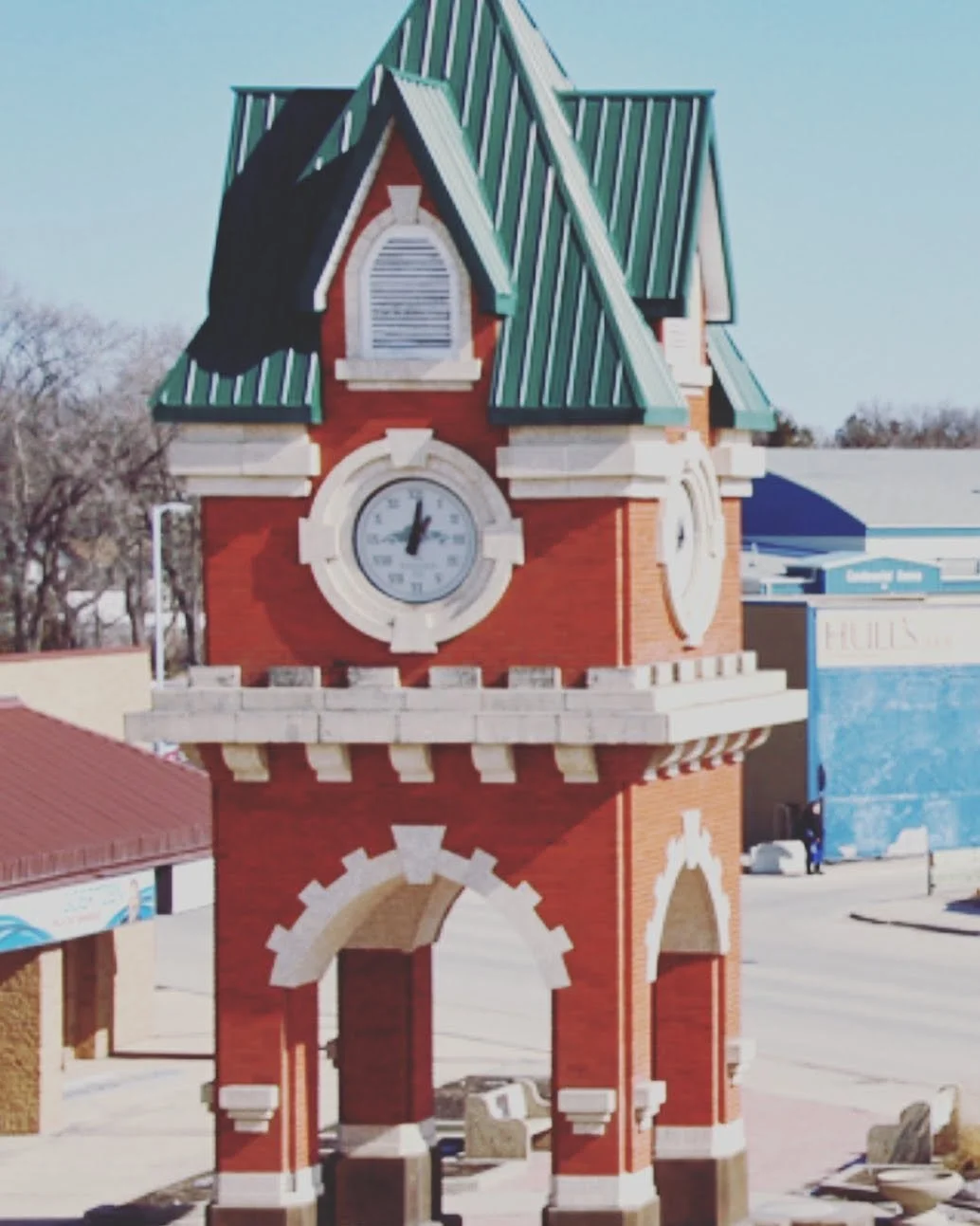 A tall clock tower with a red brick and beige stone construction, green metal roof, and a large round clock face showing 1:55, located in a small town square.