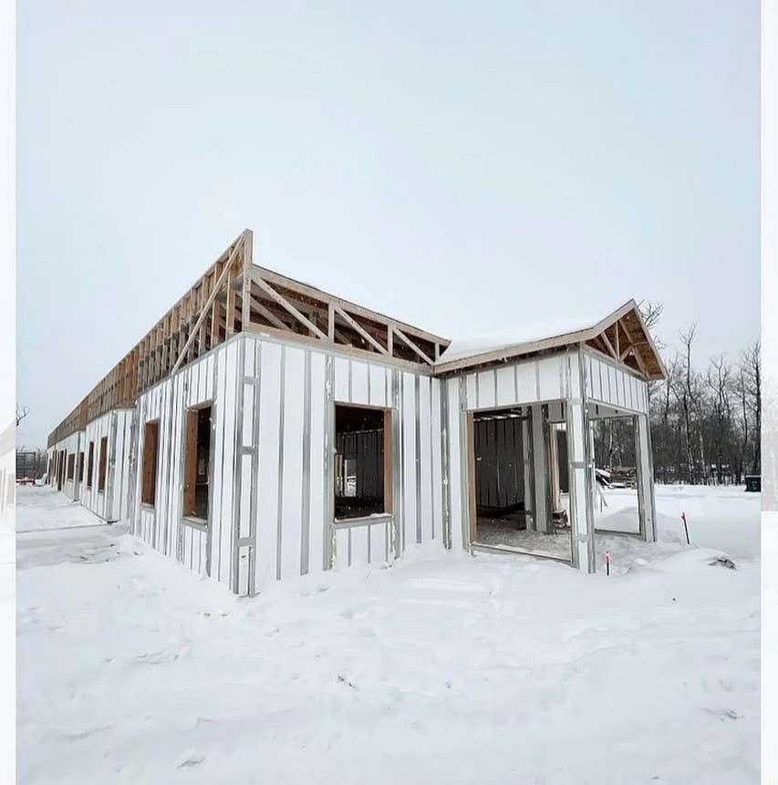A house under construction in a snowy landscape, with exterior walls and roof framing in place.