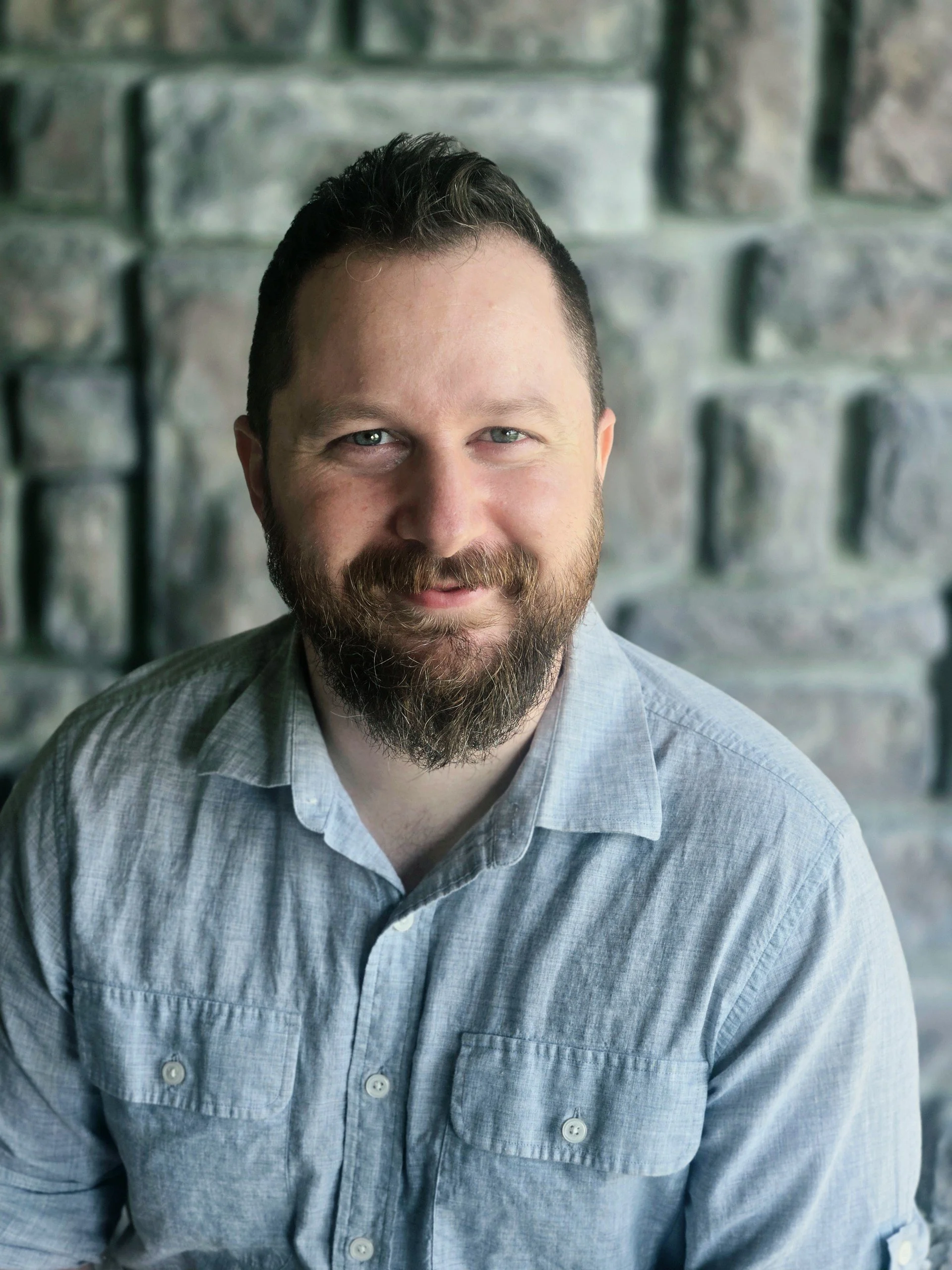 Close-up headshot of a man with dark hair, beard, and blue eyes, wearing a light blue button-down shirt, smiling slightly, in front of a blurred stone wall background.