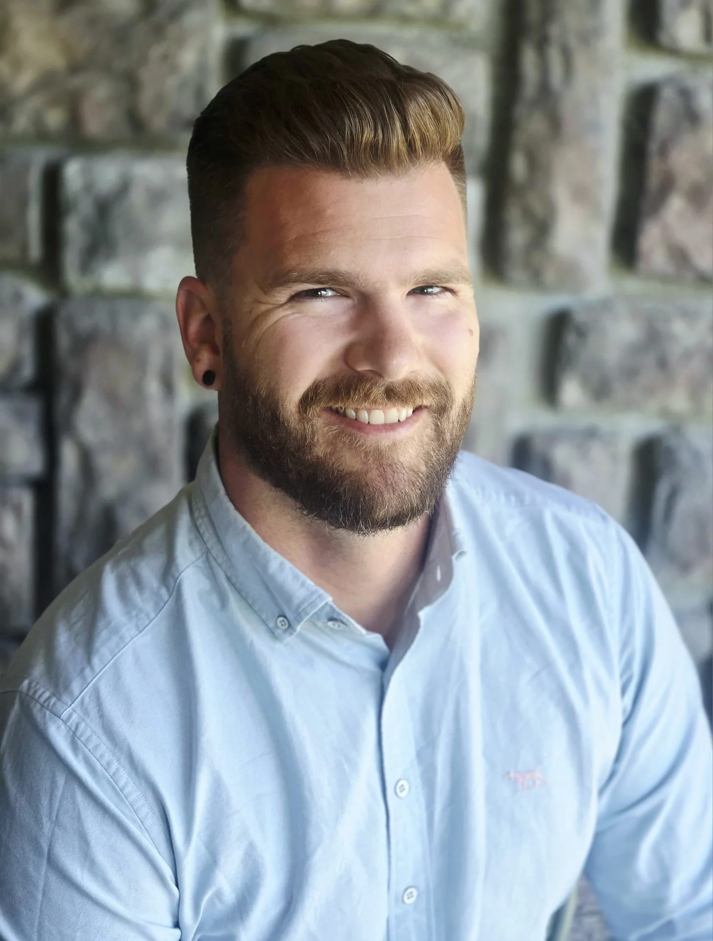 Close-up of a smiling man with light skin, styled brown hair, and a beard, wearing a light blue button-up shirt, sitting outside against a stone wall.