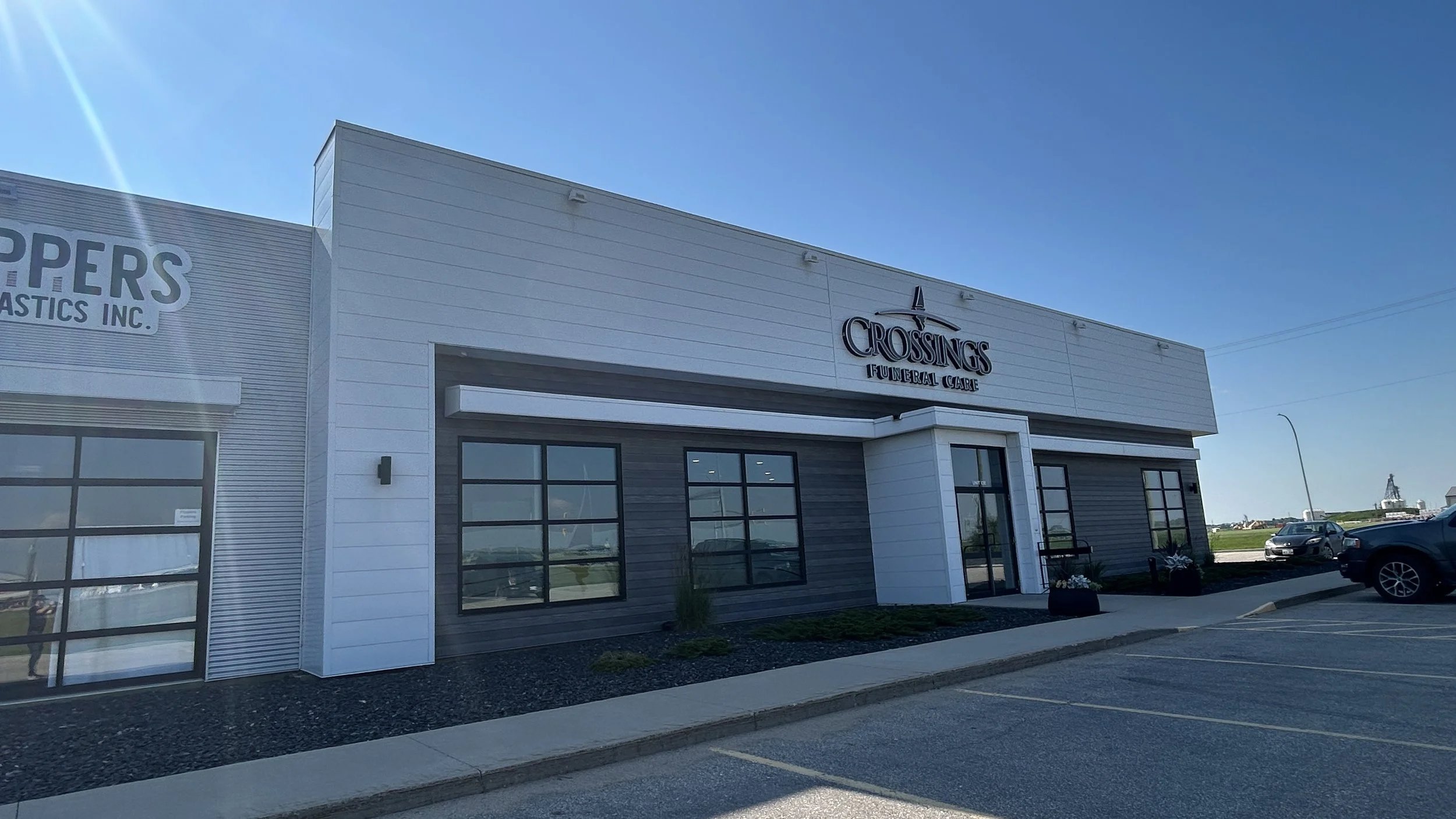 A modern white building with a sign that reads "CROSSINGS FUNERAL CARE" and an illustration of a sailboat, with parked cars in front and a clear blue sky overhead.