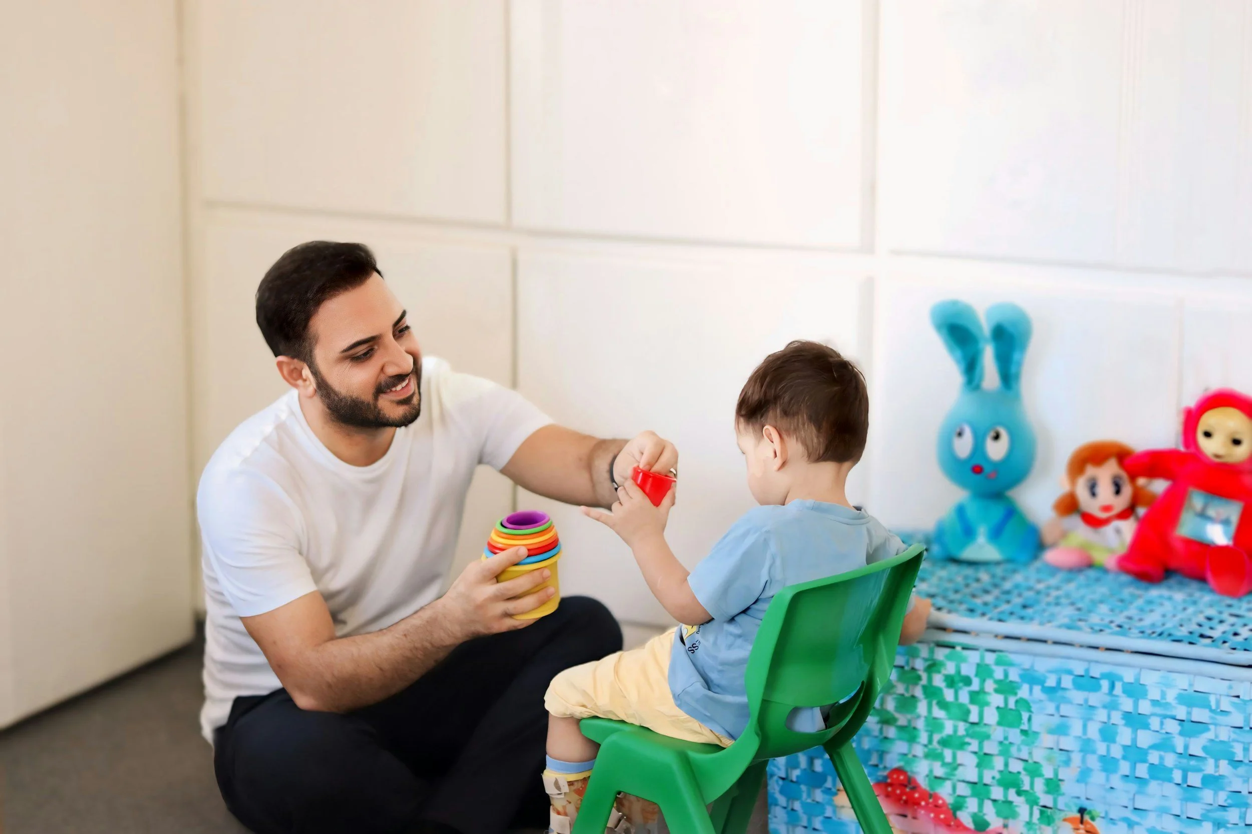 A man and young boy playing with colorful stacking cups and toys in a room decorated with toys, including a blue bunny plush and dolls.