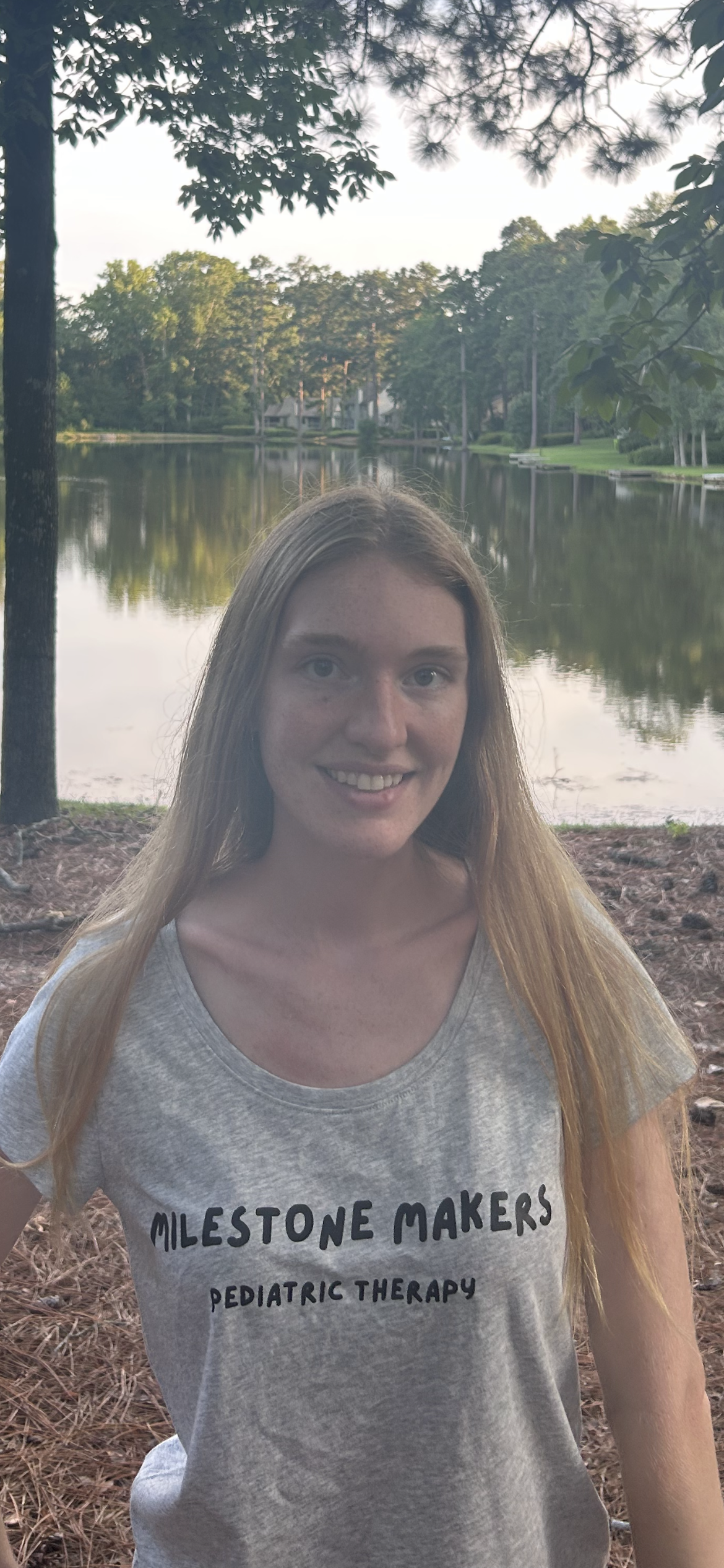 A young woman with long red hair, smiling, wearing a light gray T-shirt with black text, standing outdoors by a lake with trees and a house in the background.