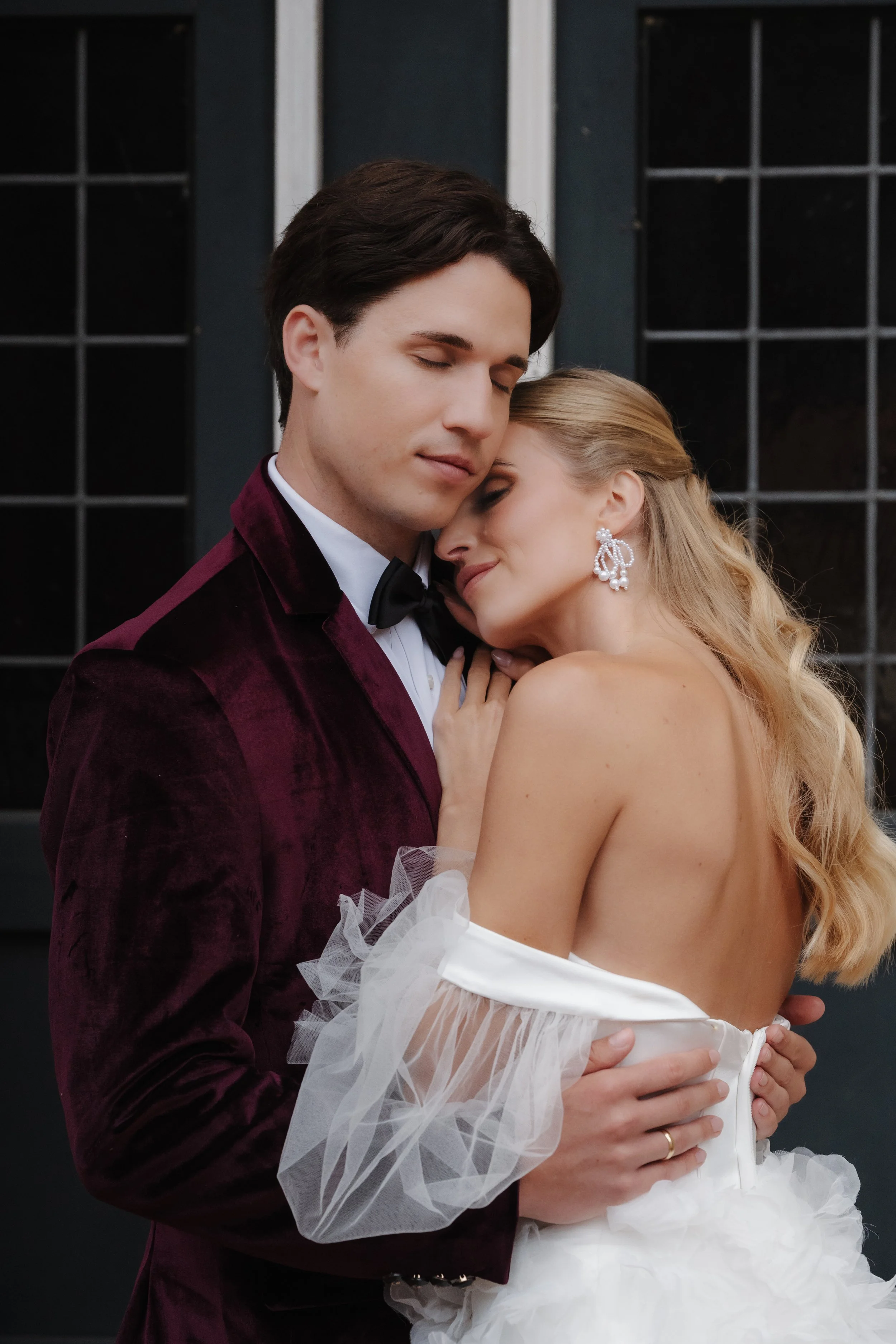 Ein verliebtes Paar in formeller Kleidung umarmt sich.
Der Mann trägt einen dunkelroten Smoking und eine Fliege, die Frau trägt ein weißes Hochzeitss Kleid und große Ohrringe.