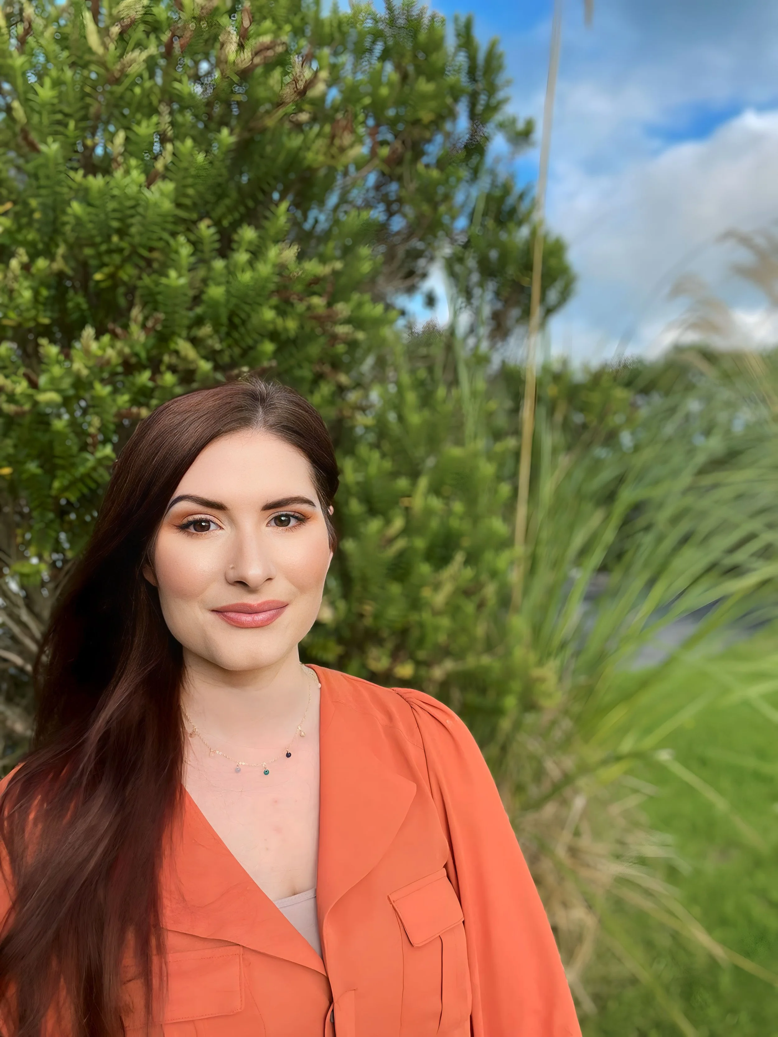 Close-up of a woman with long brown hair, wearing an orange blazer and a delicate necklace, standing outdoors with lush green trees and blue sky in the background.