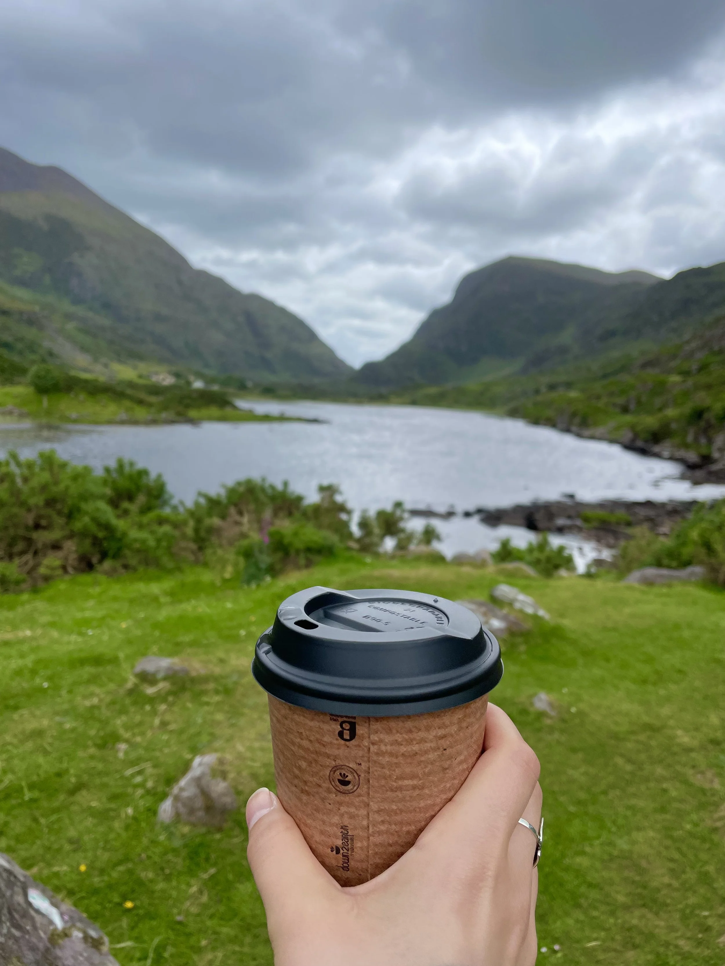 A hand holding a coffee cup in front of a scenic landscape with a river, green hills, and mountains under a cloudy sky.