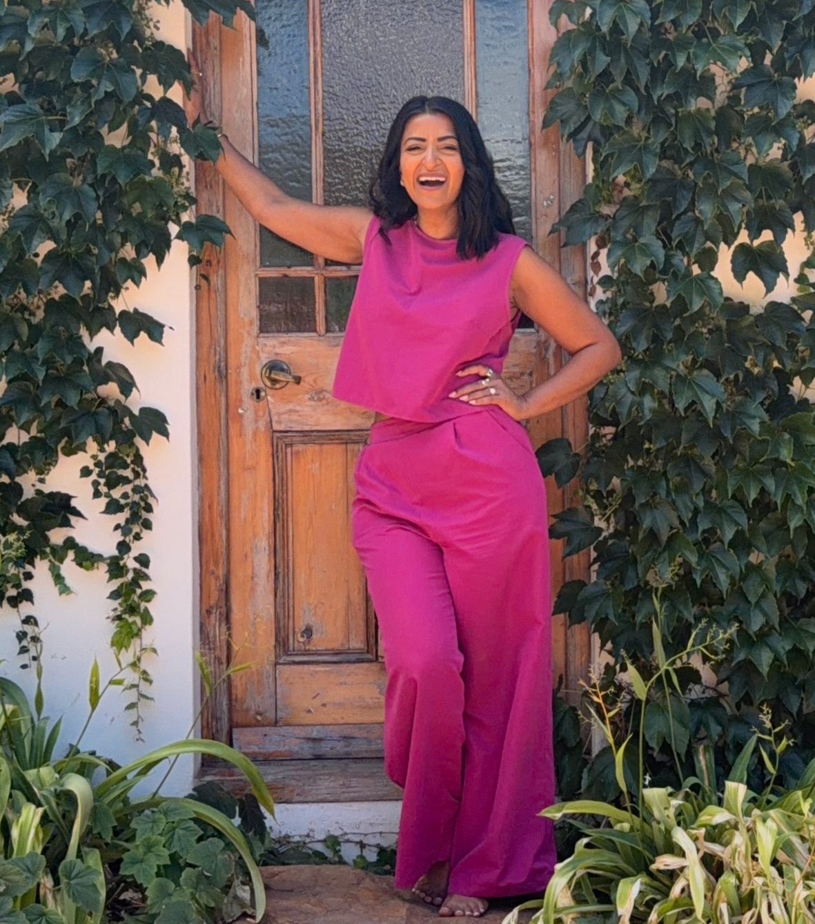 A woman standing outdoors in front of a wooden door, wearing a pink sleeveless top and matching wide-leg pants, smiling with one hand on her hip and the other resting on the door frame, surrounded by green foliage.