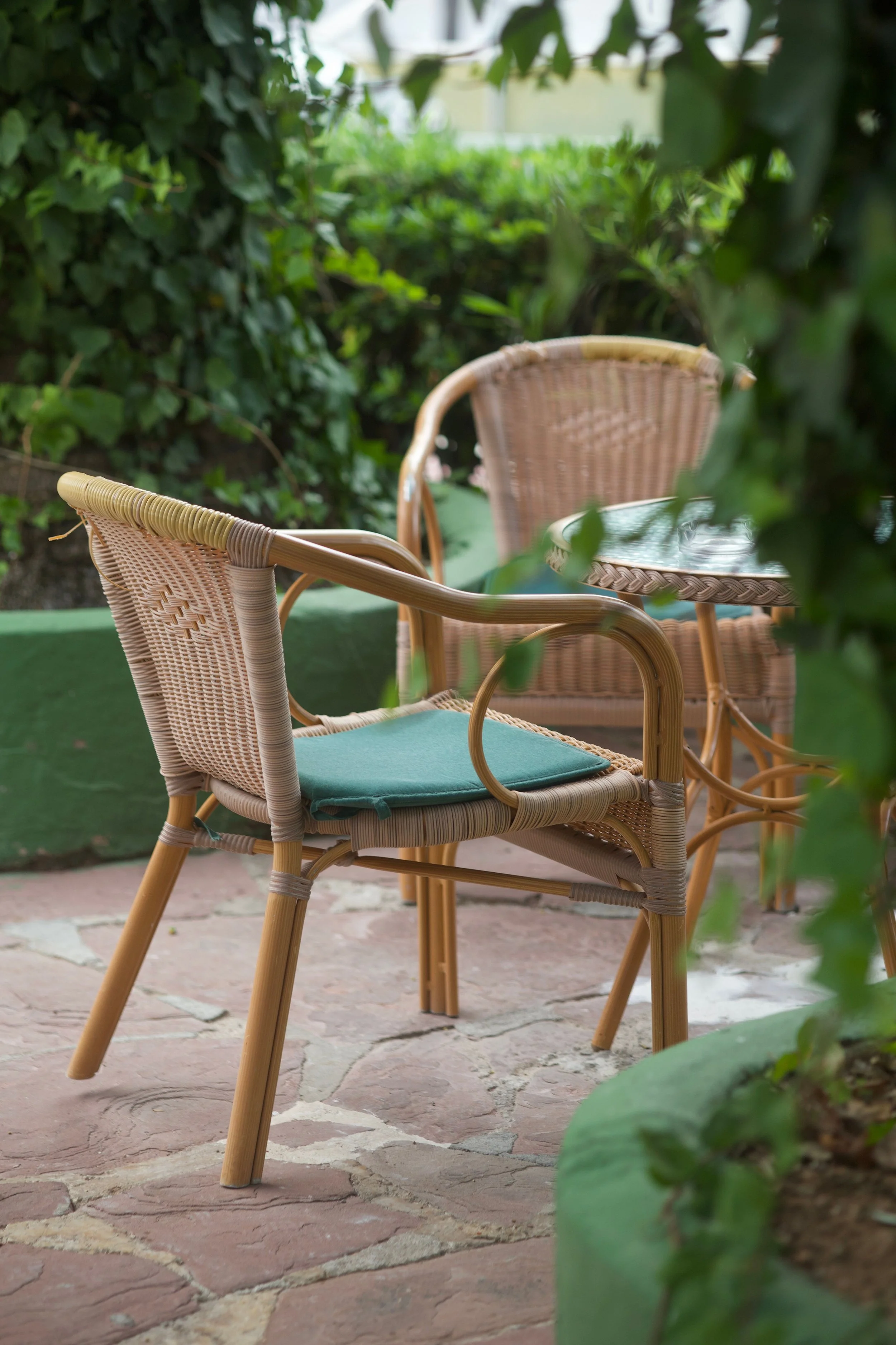 Two wicker chairs with blue cushions around a glass-topped wicker table outdoors, surrounded by green plants on a stone patio.