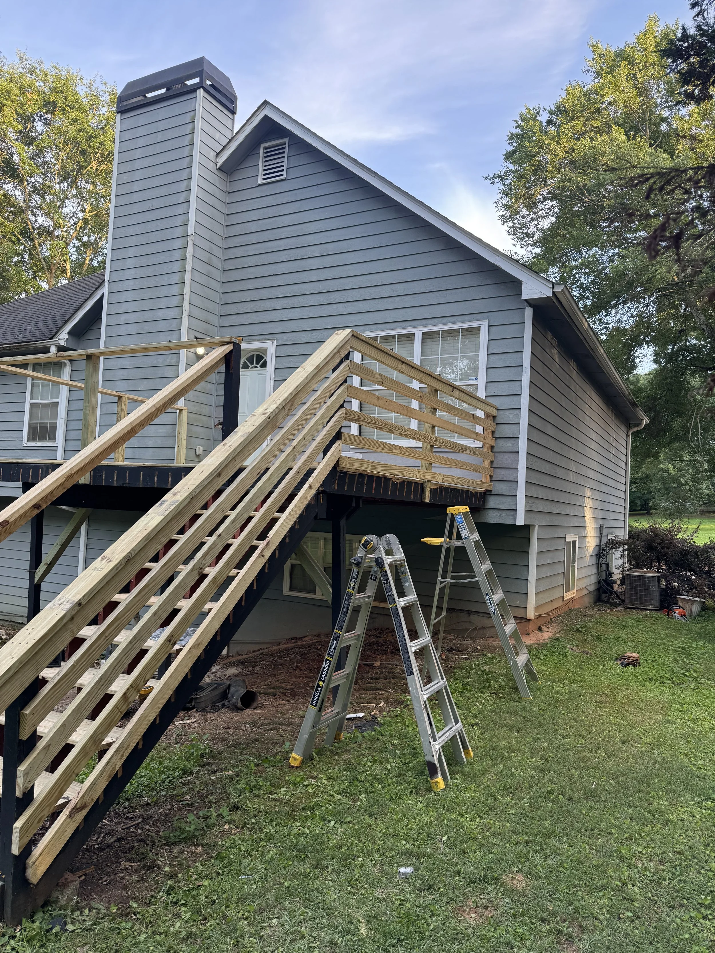 House with gray wooden siding, a staircase leading up to a door, and ladders in the yard. Construction or renovation work in progress on the house.