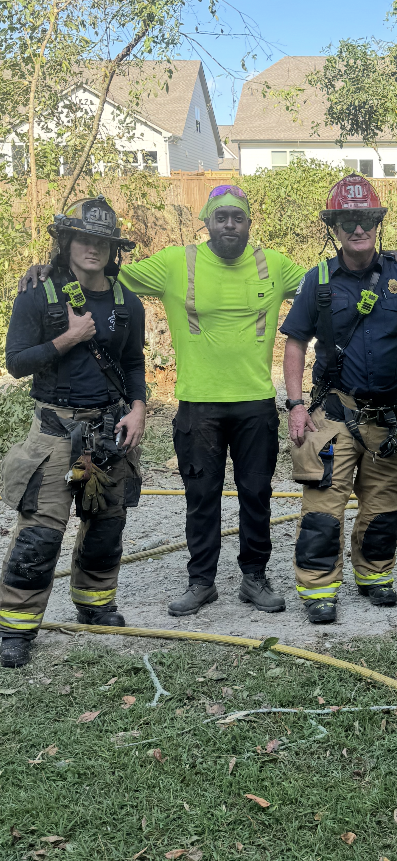 Three emergency responders standing outdoors on a dirt surface, with two house yards and trees in the background. They are wearing helmets and safety gear, and appear to have been working at a site, possibly a fire or rescue operation.