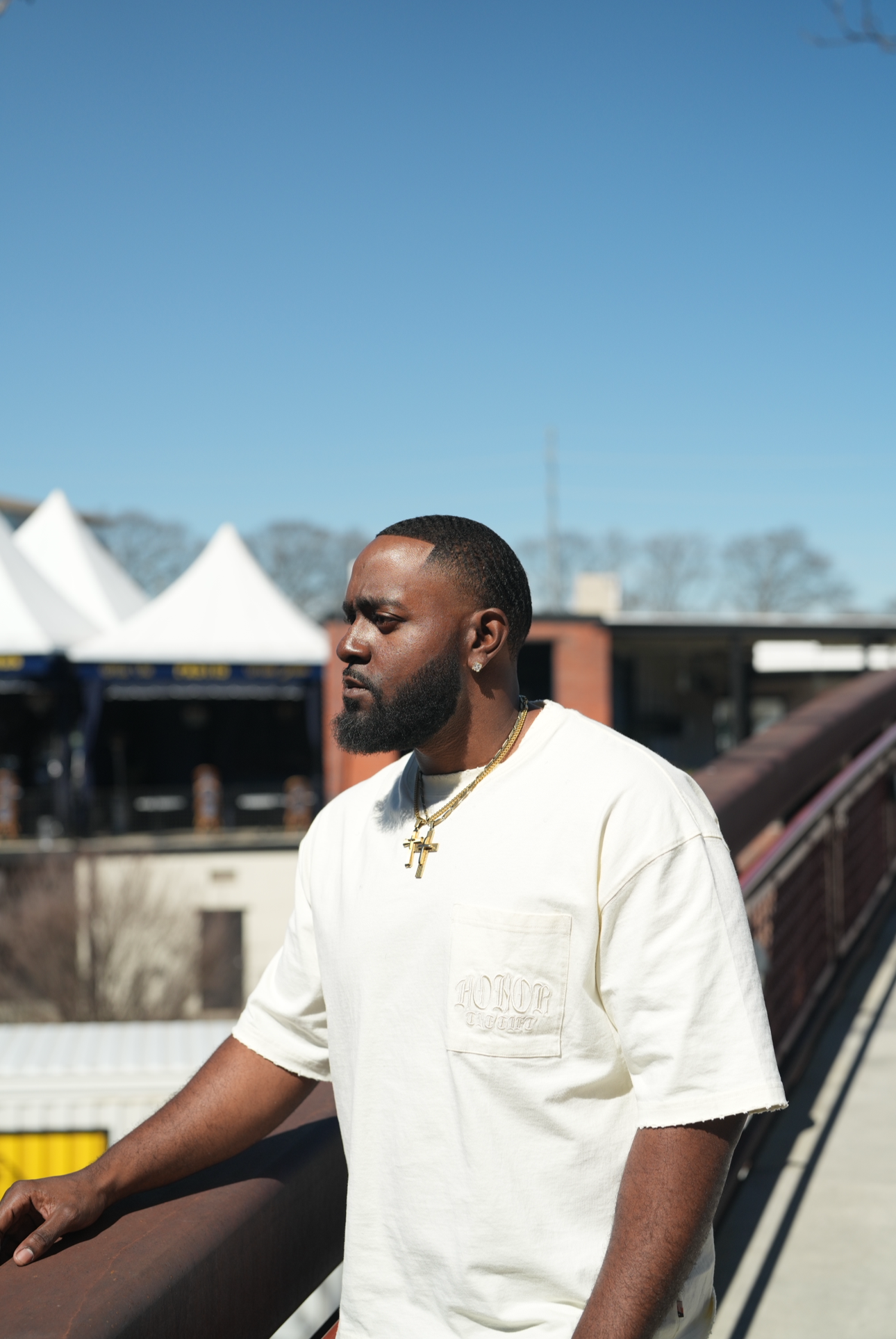 A man with a beard and short hair, wearing a beige T-shirt and gold jewelry, stands outdoors on a rooftop with a cityscape in the background under a clear blue sky.