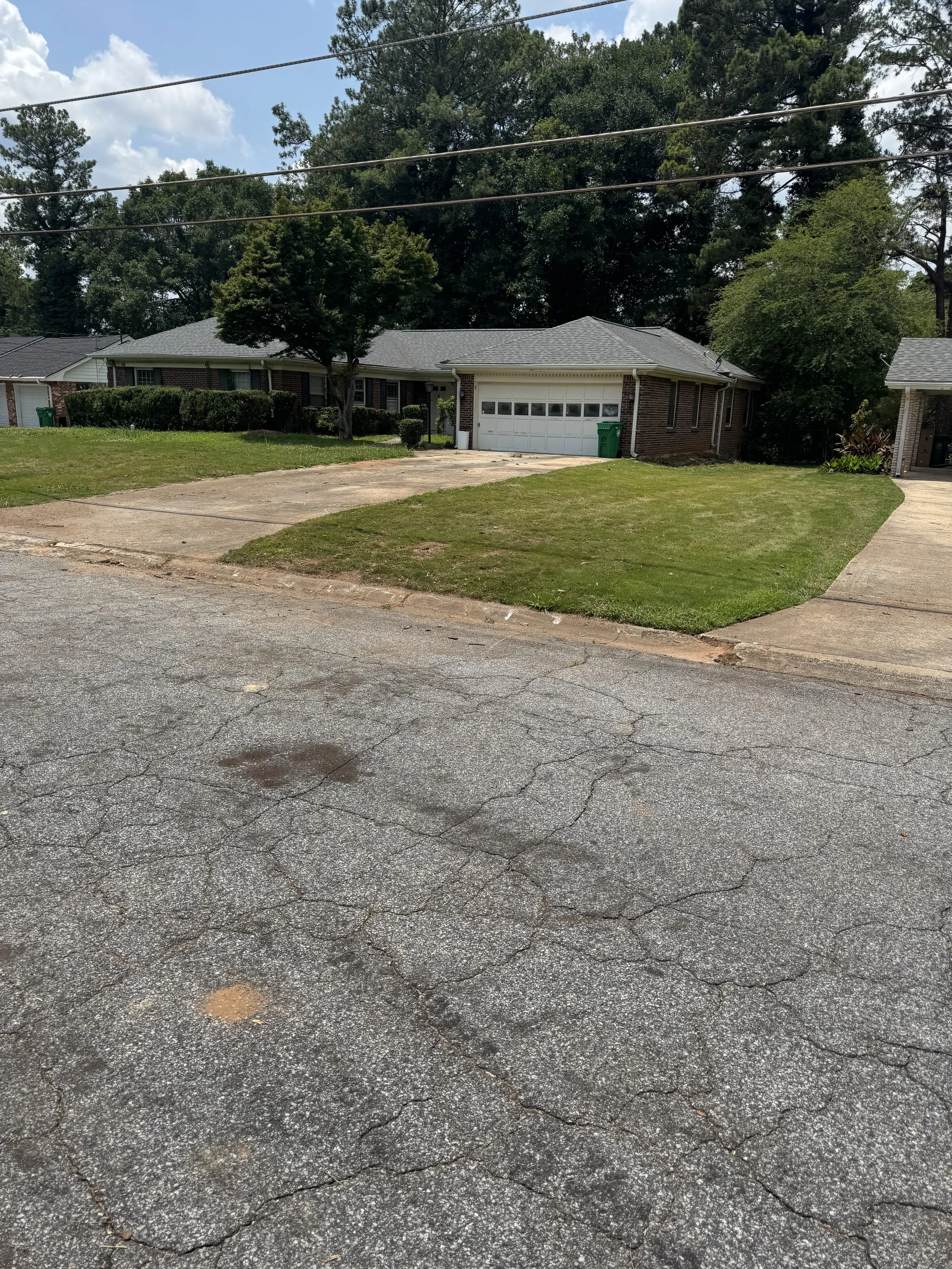 A residential neighborhood with a one-story brick house, a driveway, a front lawn, and trees in the background. The pavement in the foreground is cracked.