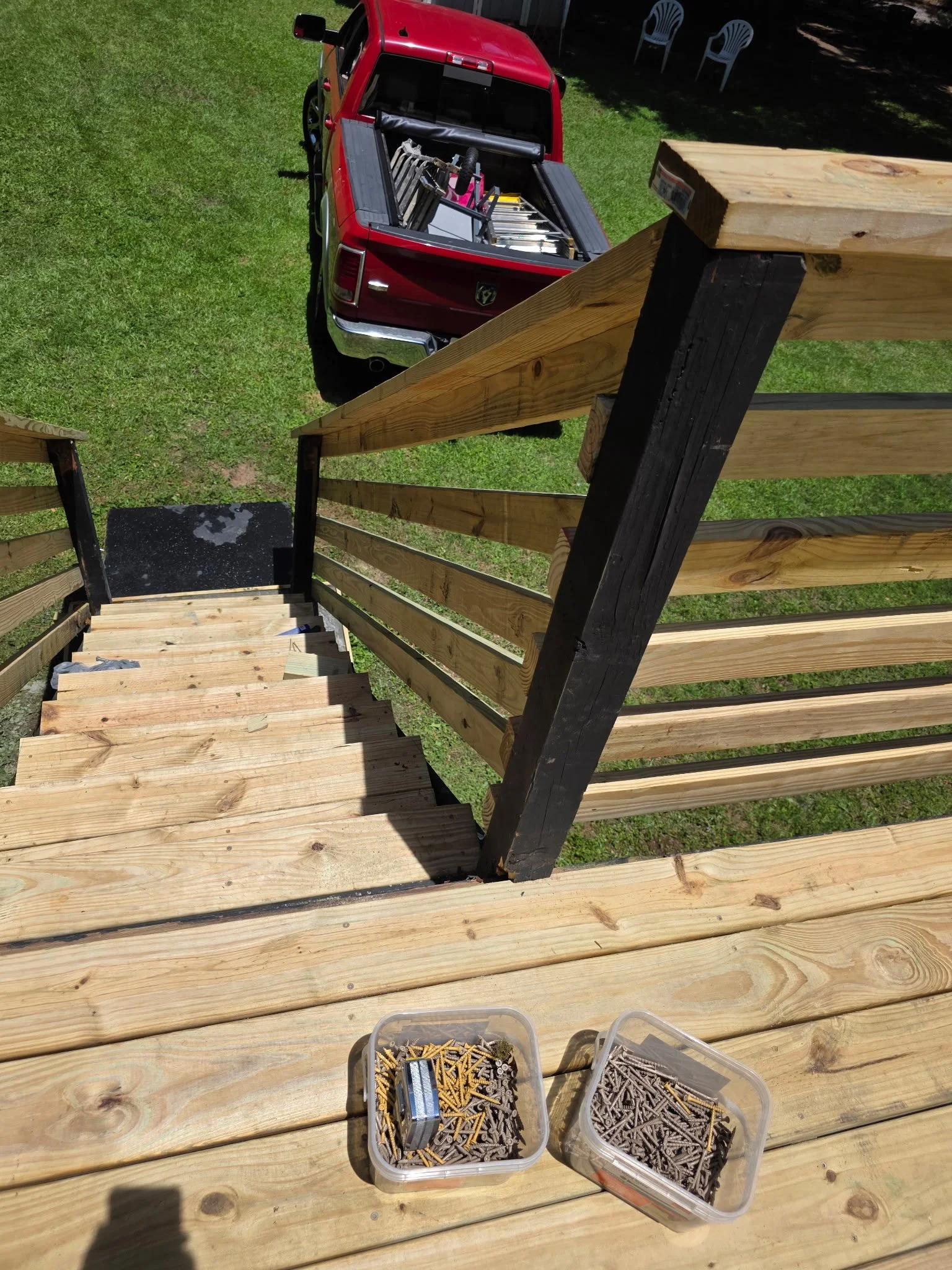 Wooden stairs leading down to a grassy yard with a red pickup truck parked on the grass. Two plastic containers filled with nails are on the stairs, and a toolbox is visible at the bottom of the stairs.