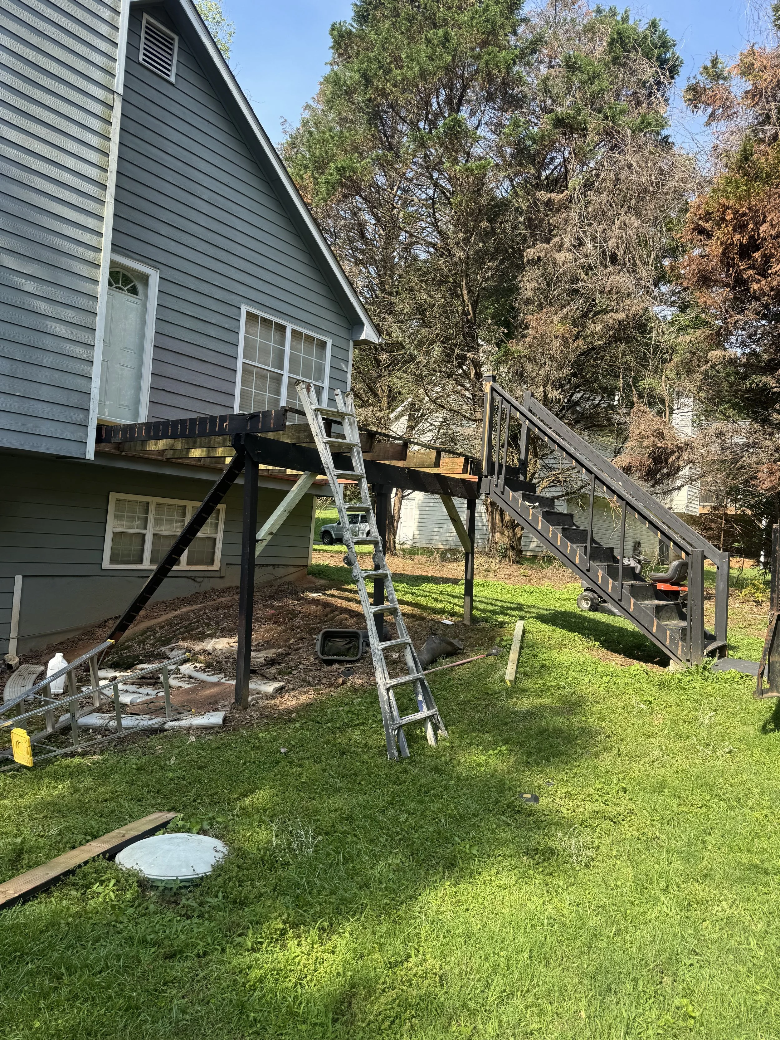 A house with blue siding and white trim, with a gray door and two windows. An outdoor deck is being built or repaired, with a metal ladder and a black staircase leading to it. There are construction tools and materials around, with a grassy yard and trees in the background.