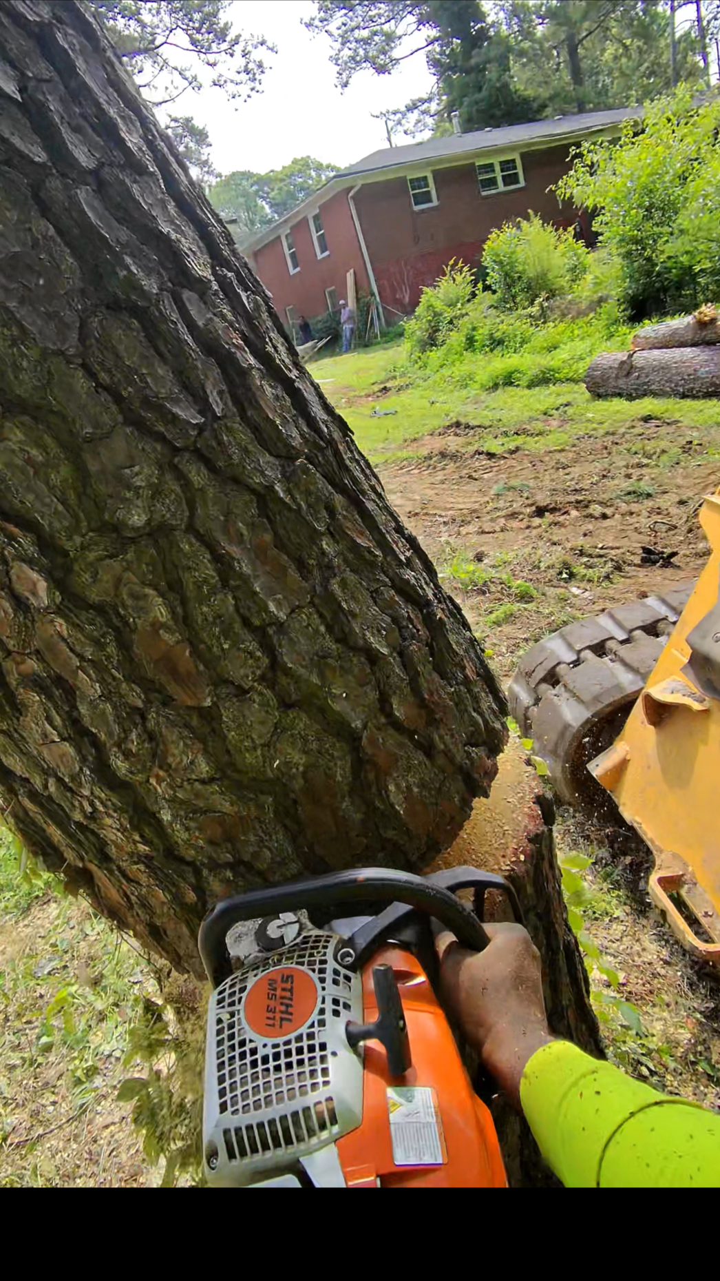 A person using a chainsaw to cut down a large tree trunk in a backyard. The chainsaw is orange and black, and the person is wearing a bright yellow safety shirt. The background shows a house, green trees, and shrubs.