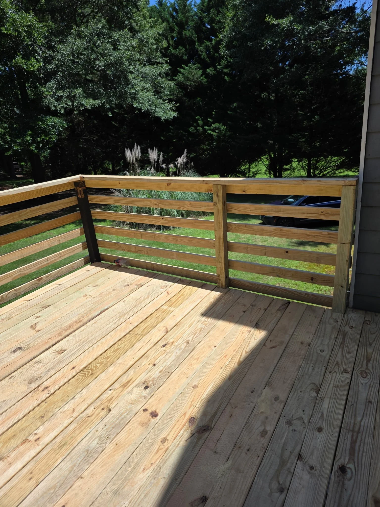 View of a newly built wooden deck with a railing, overlooking a green yard and dense trees. Part of a house's exterior wall is visible on the right side.